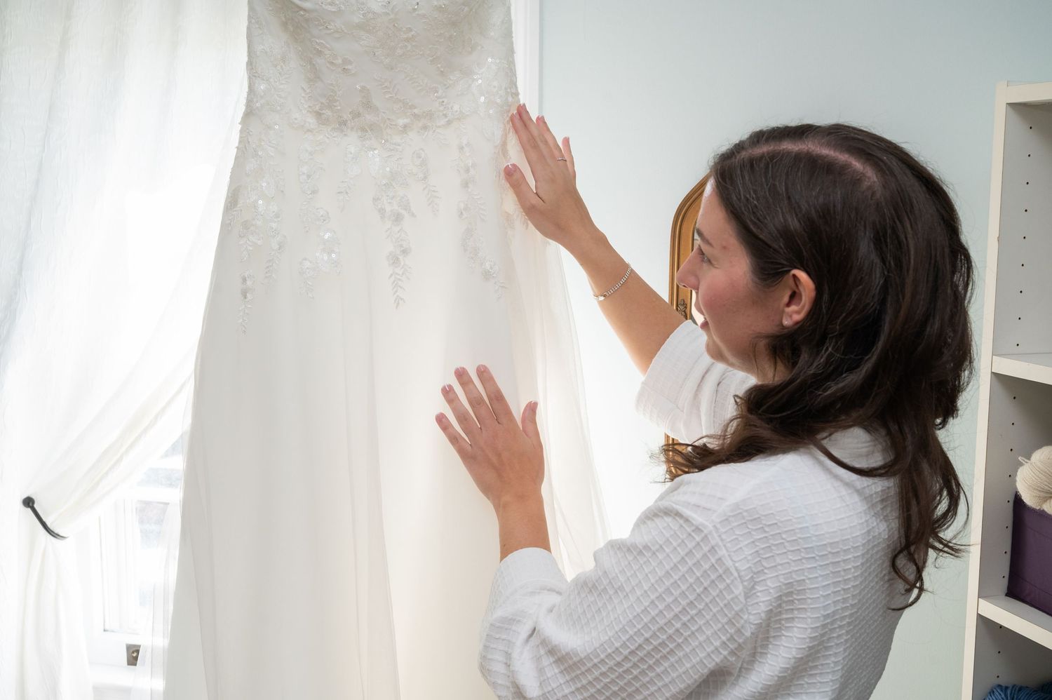 Bride in white robe admiring her lace wedding dress hanging by a bright window.