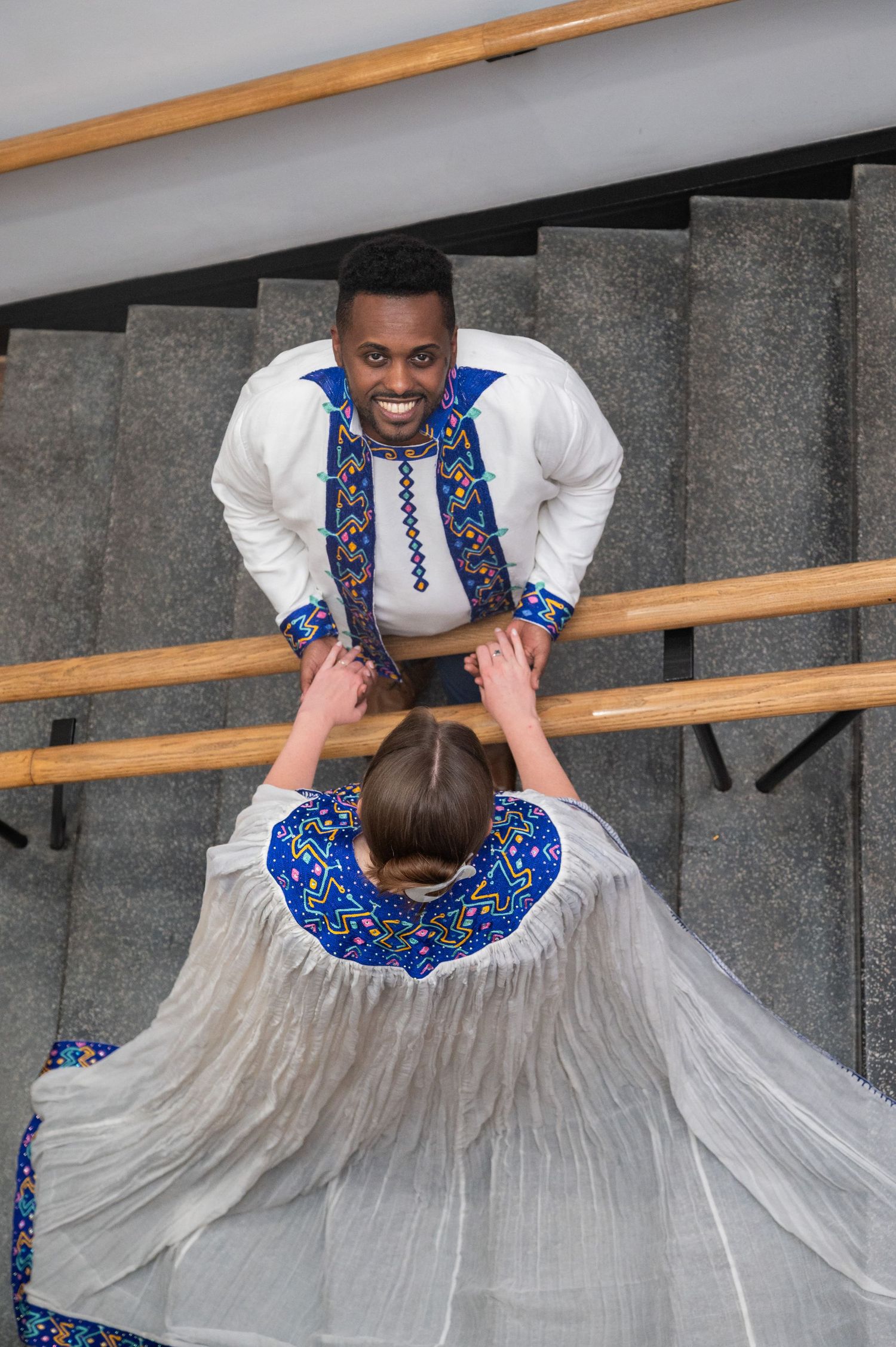 Couple in matching blue and white African traditional attire holding hands on stairs, viewed from above.