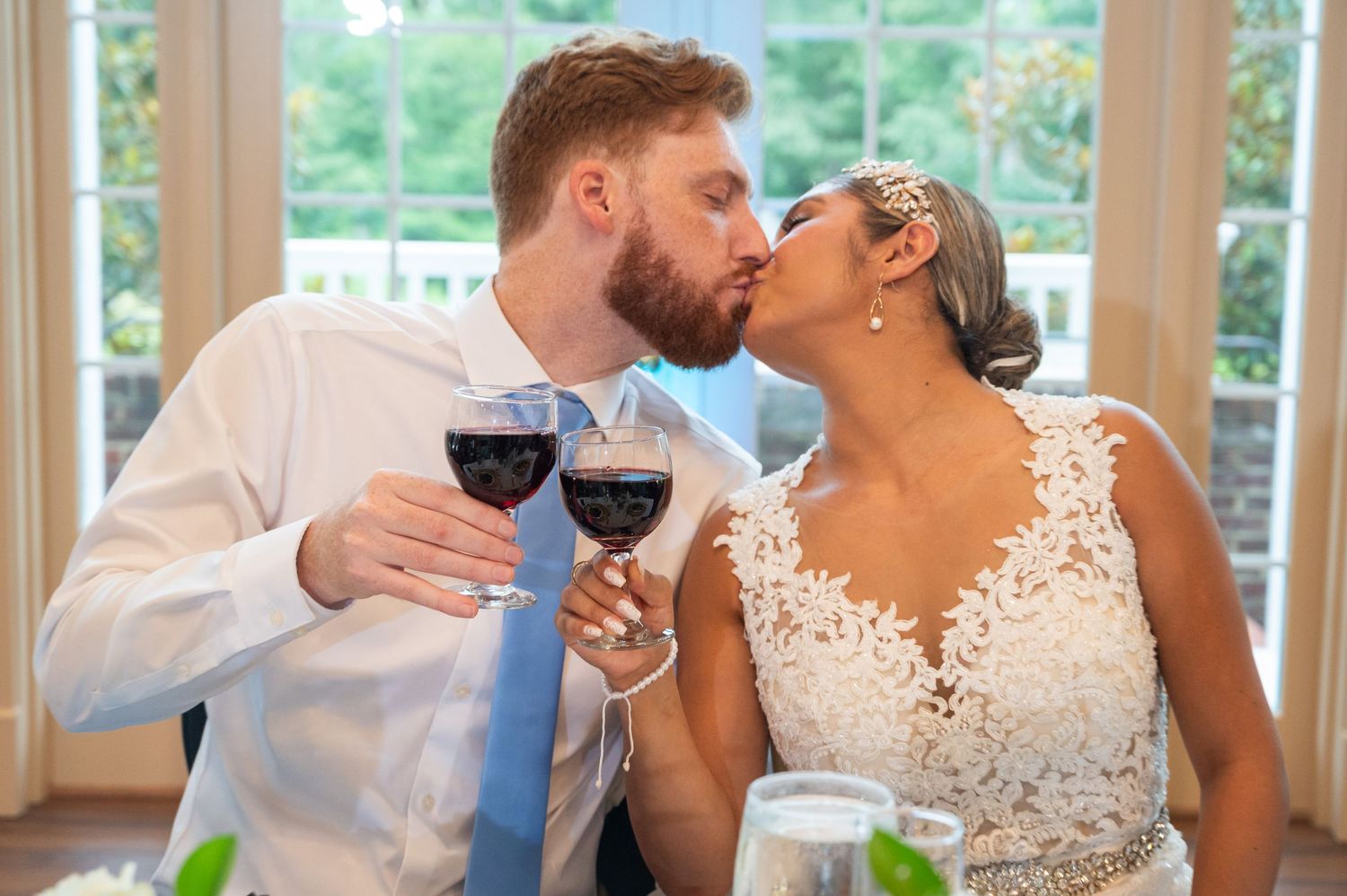 Newlyweds share a kiss while toasting with red wine glasses at their wedding reception indoors.