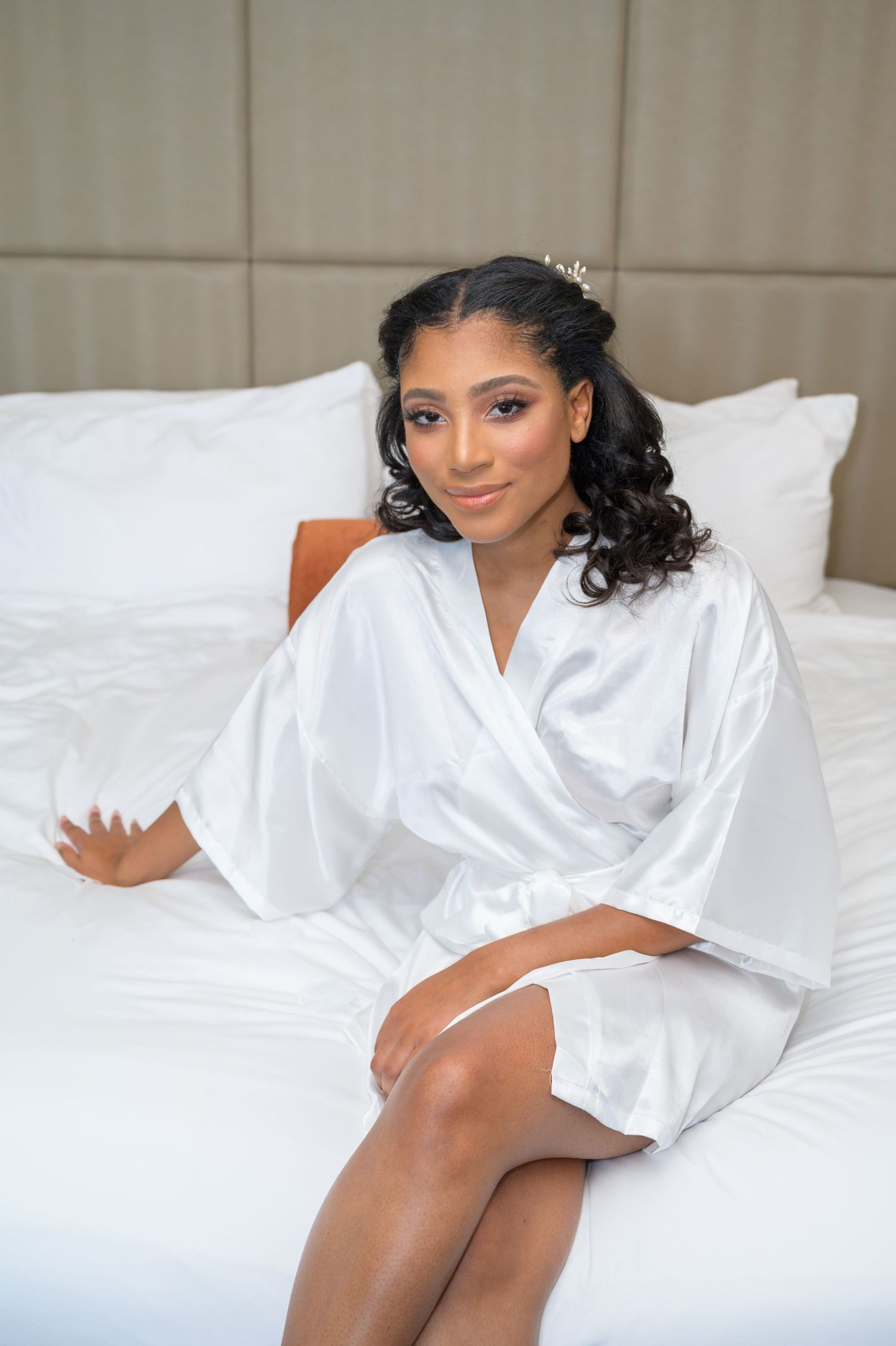Elegant bride in white satin robe sits on hotel bed with curly hair adorned with accessories.