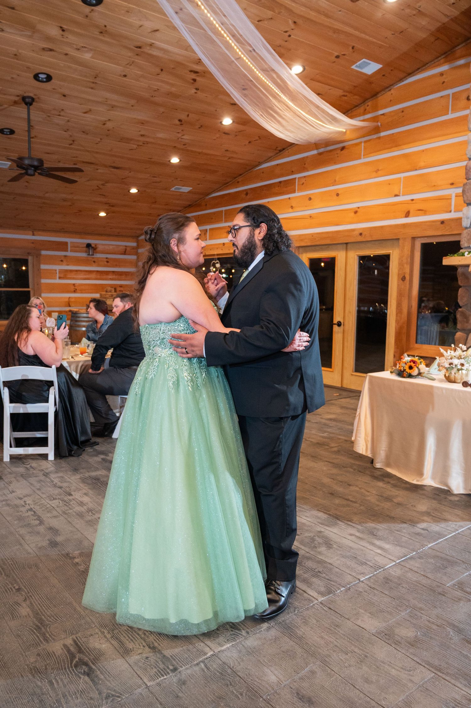 Newlyweds share first dance in rustic log cabin wedding venue with guests seated in background.