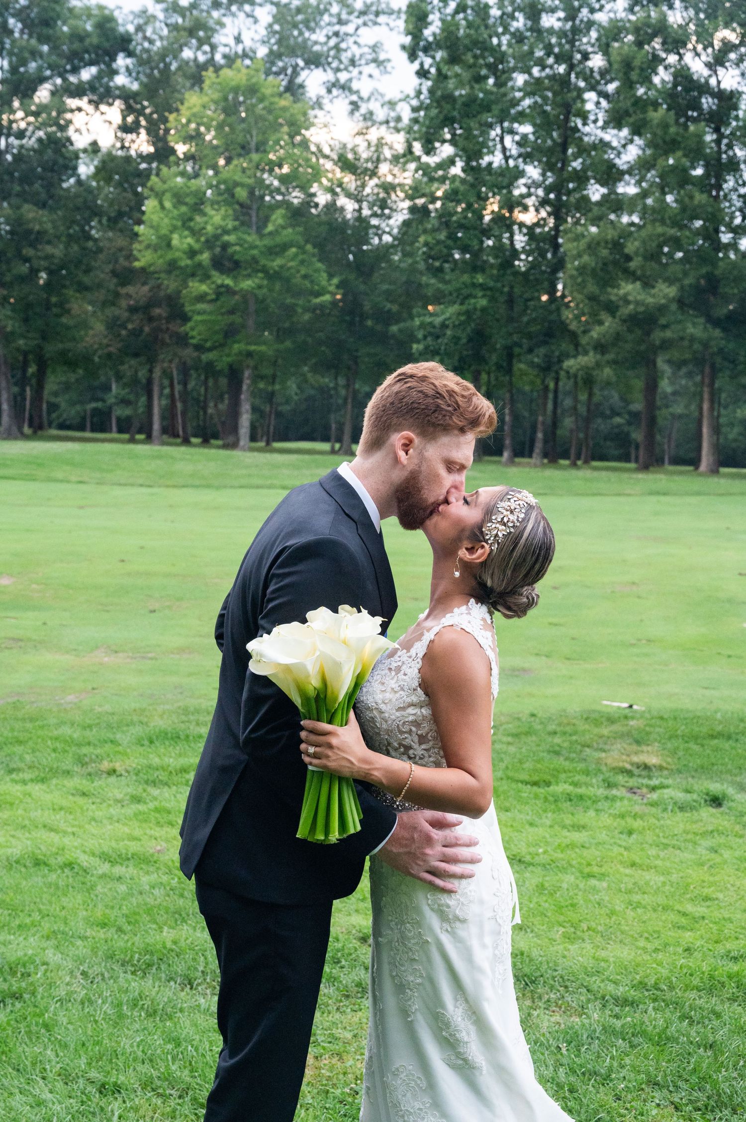 Bride and groom share a romantic kiss on a lush green lawn, bride holding white calla lily bouquet.