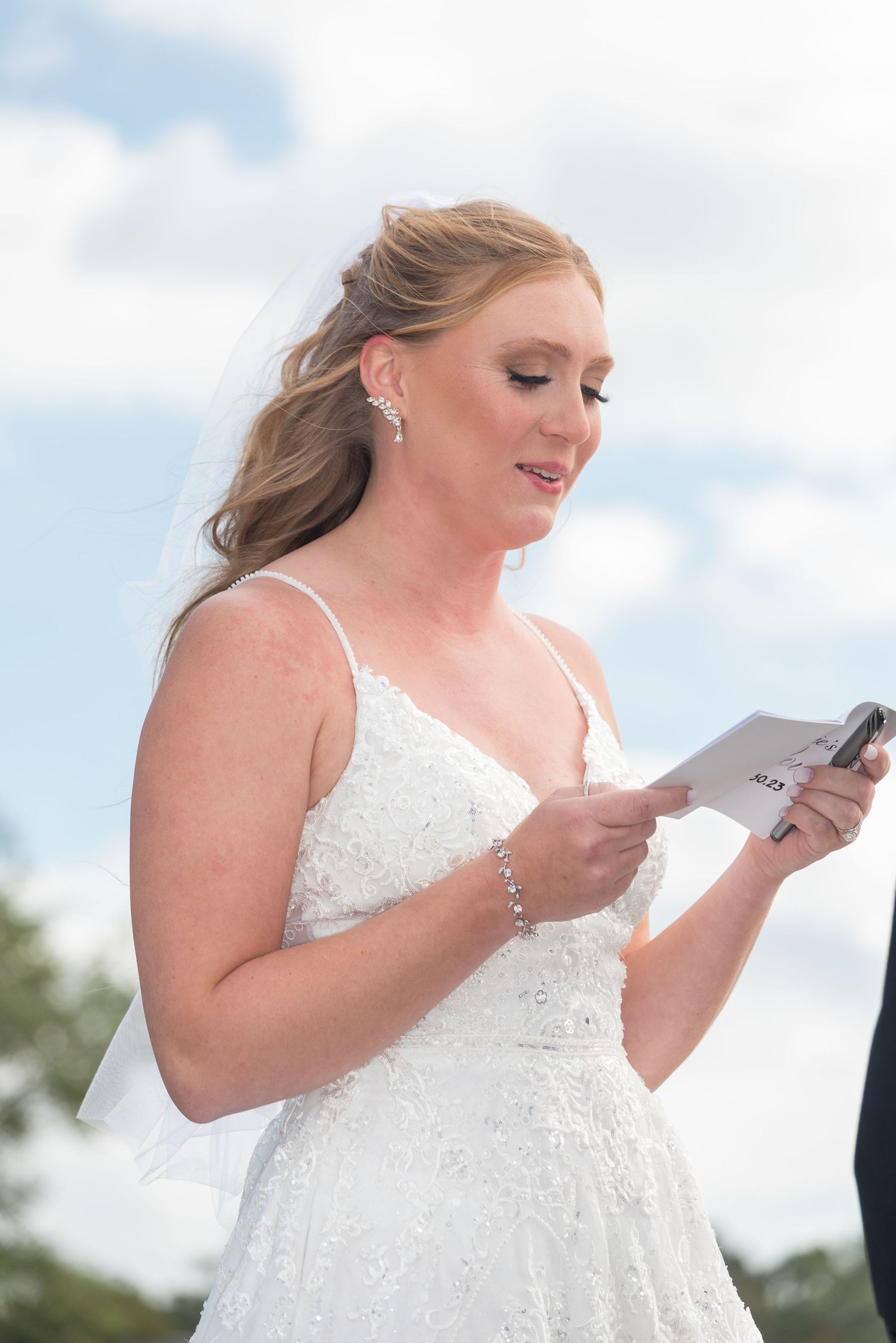 Beautiful bride in lace wedding gown reading vows from card during outdoor wedding ceremony.
