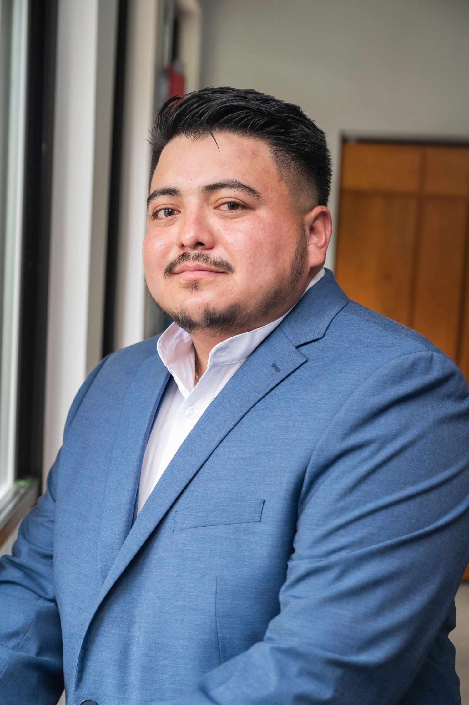 Man in blue suit with white dress shirt poses for formal portrait near window.