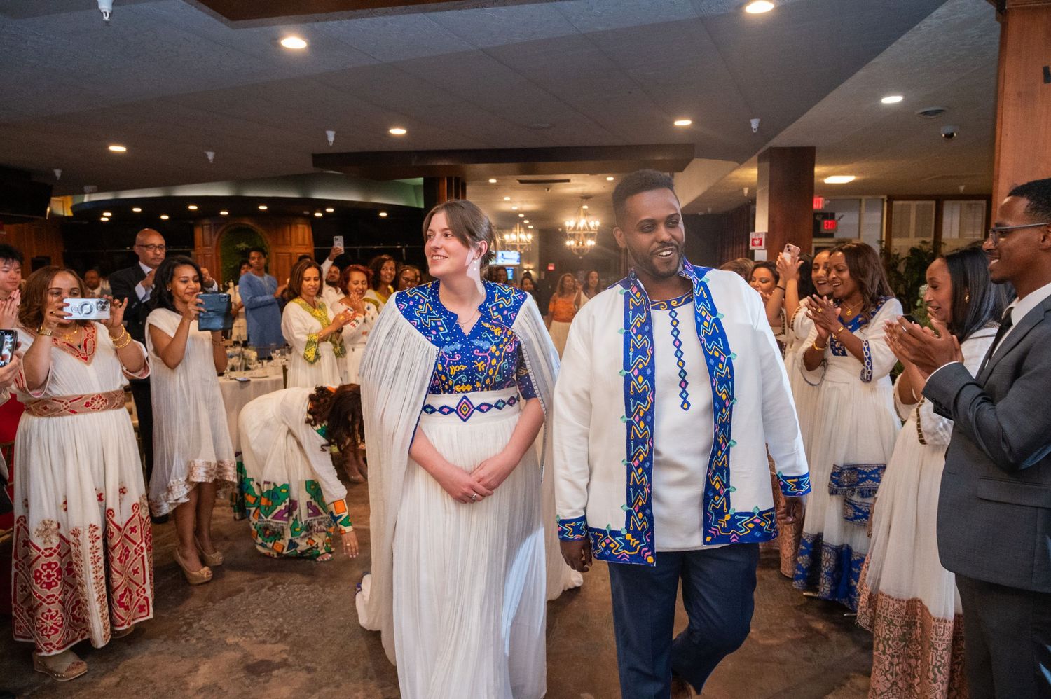 Couple in traditional Ethiopian white and blue cultural attire entering wedding reception hall.