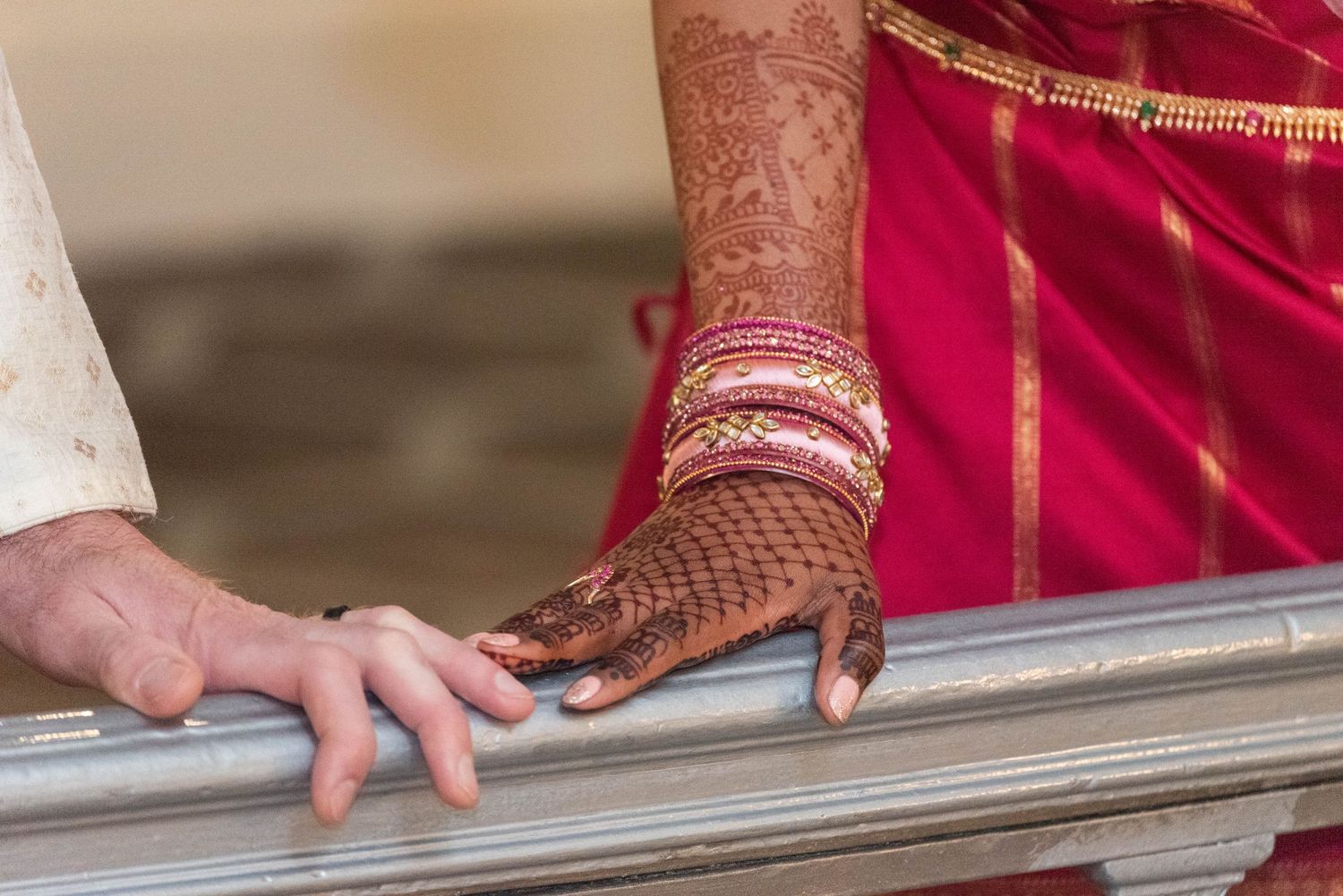 Close-up of Indian bride's henna-adorned hand with red and gold bangles resting on a railing beside groom's hand.