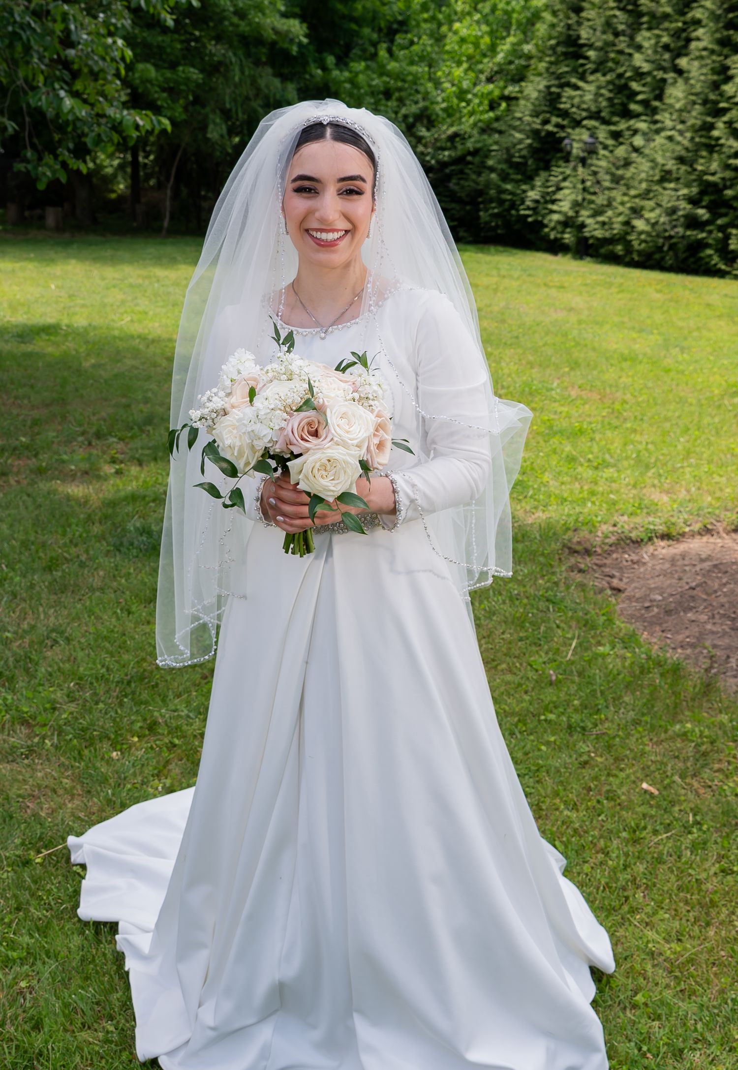 Smiling bride in white long-sleeve wedding gown and veil holding bouquet of blush roses outdoors.