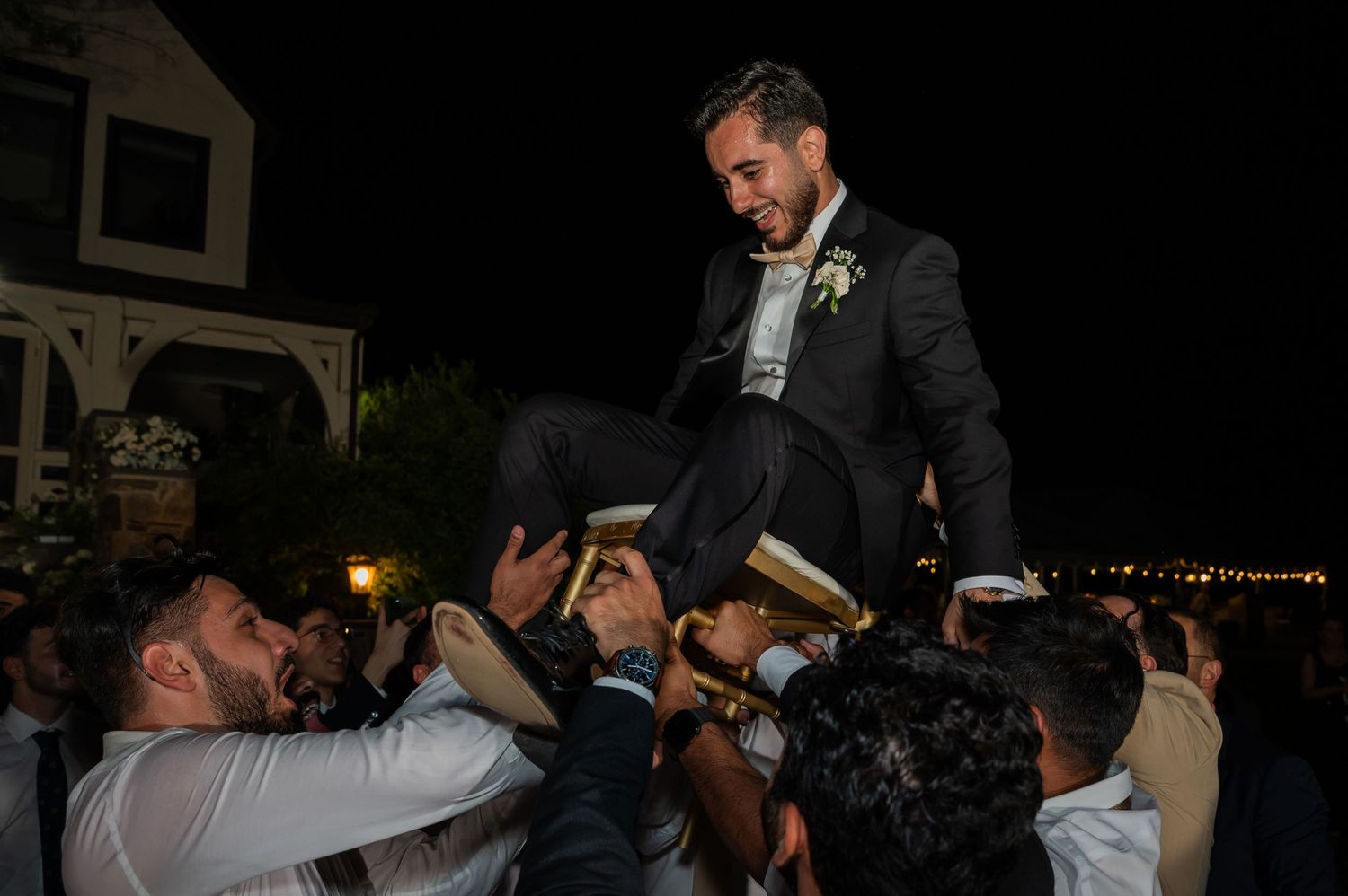 Groom in black tuxedo lifted by guests during outdoor wedding celebration at night.