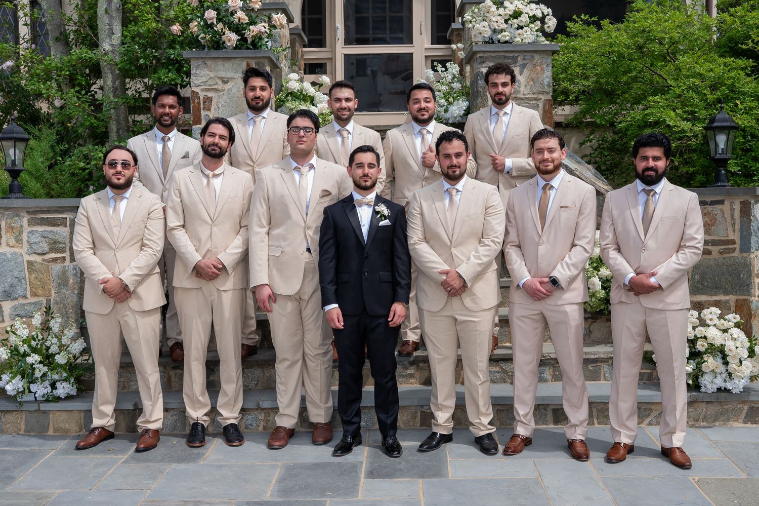 Groom in black suit poses with groomsmen in beige suits outside a stone building with white flowers.