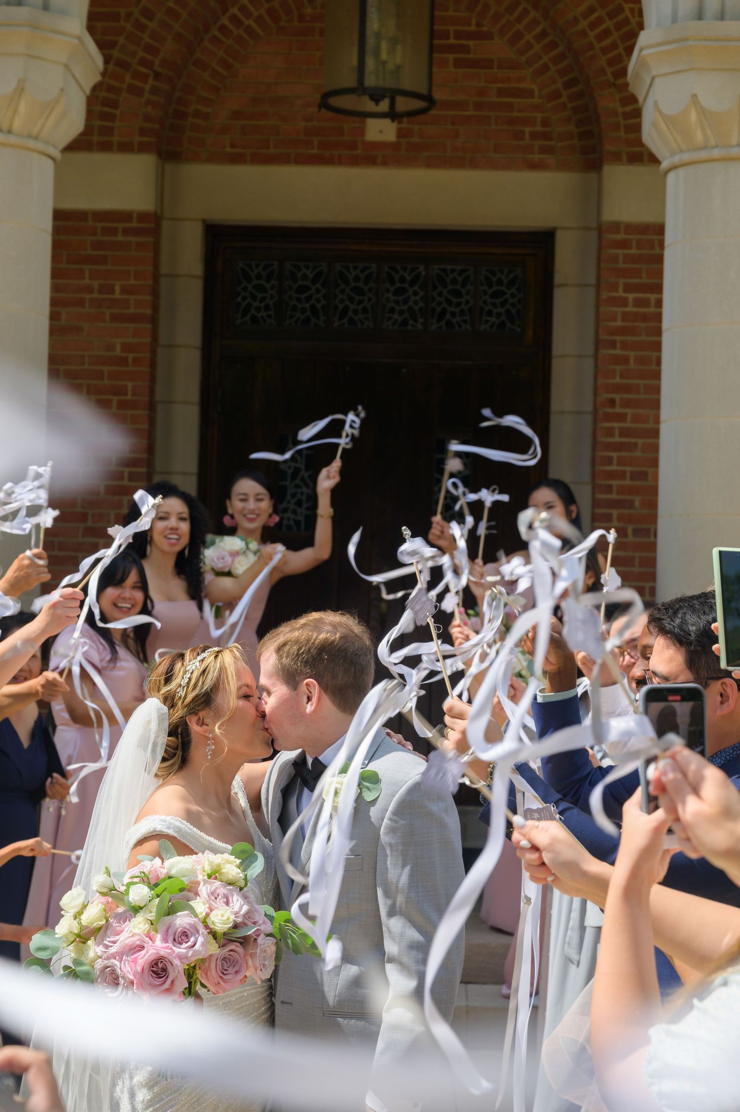 Newlyweds kiss as guests wave white ribbon wands outside brick church after wedding ceremony.