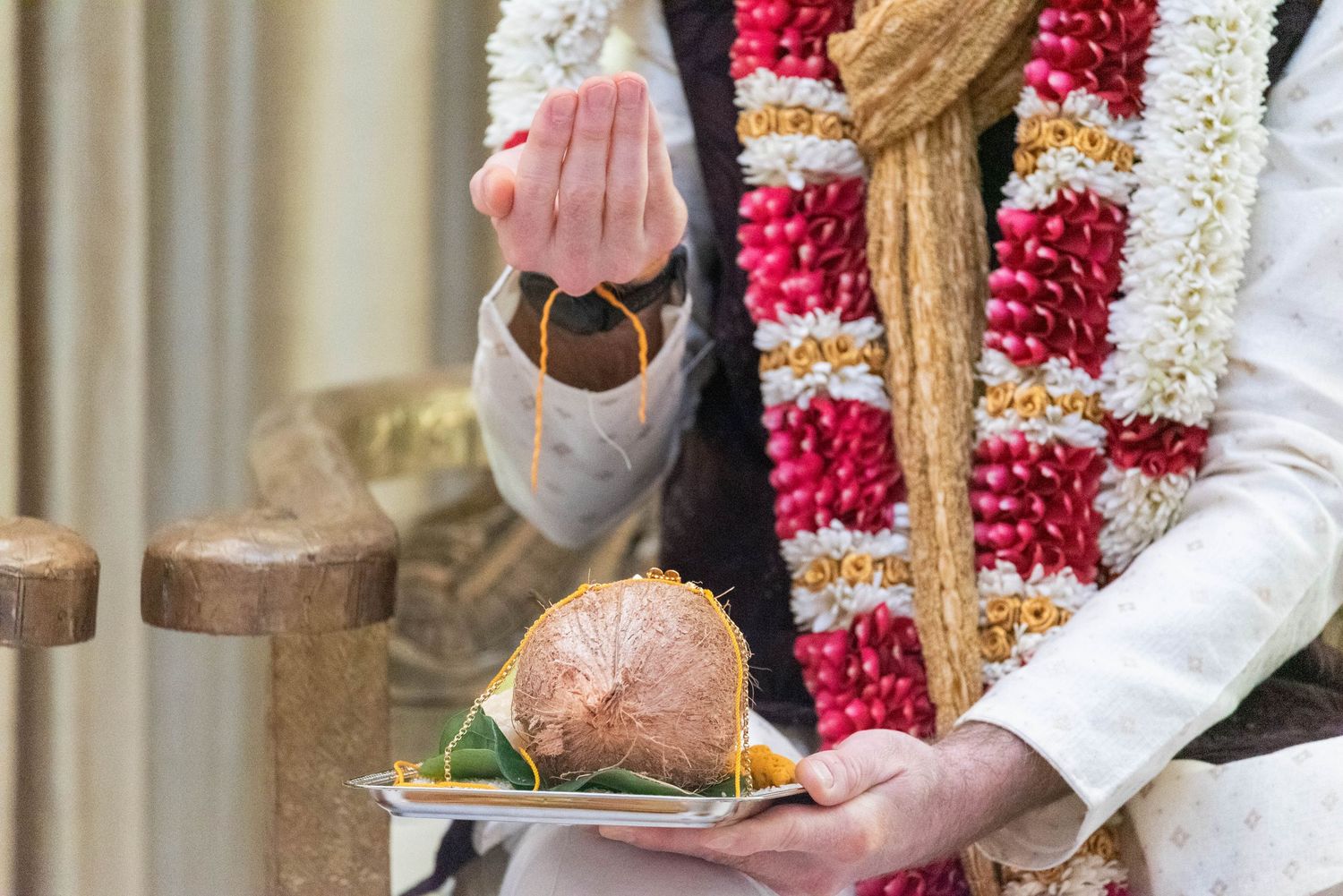 Man in traditional Indian wedding attire holding a coconut on a plate and a small pot during a Hindu ceremony.