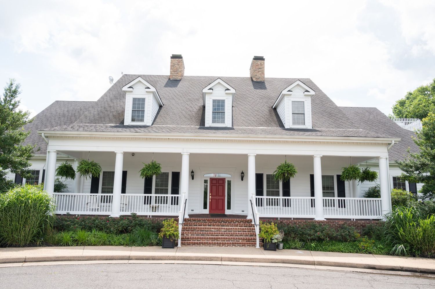 White Southern colonial home with wrap-around porch, dormer windows, red front door, and brick steps surrounded by lush greenery.