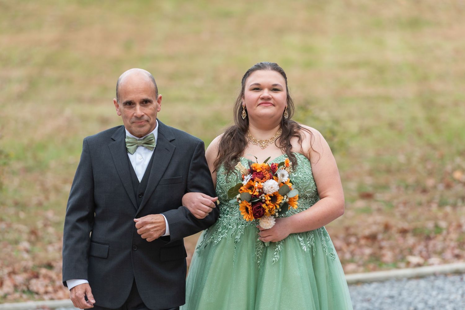 A bride in a green gown holds a fall bouquet while walking with a man in a tuxedo at an outdoor wedding.