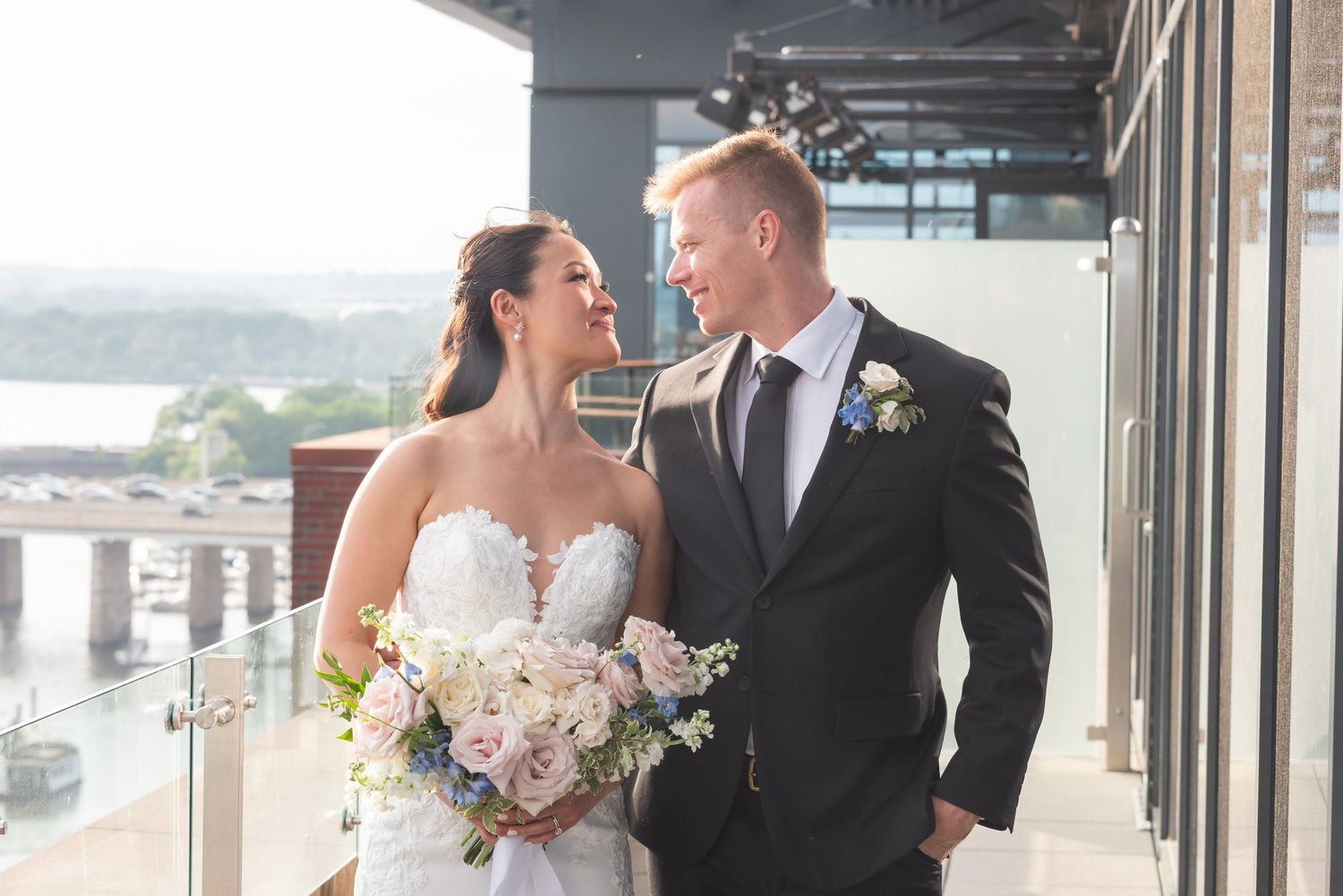 Elegant bride and groom posing on waterfront venue balcony with lush bouquet of pastel flowers.