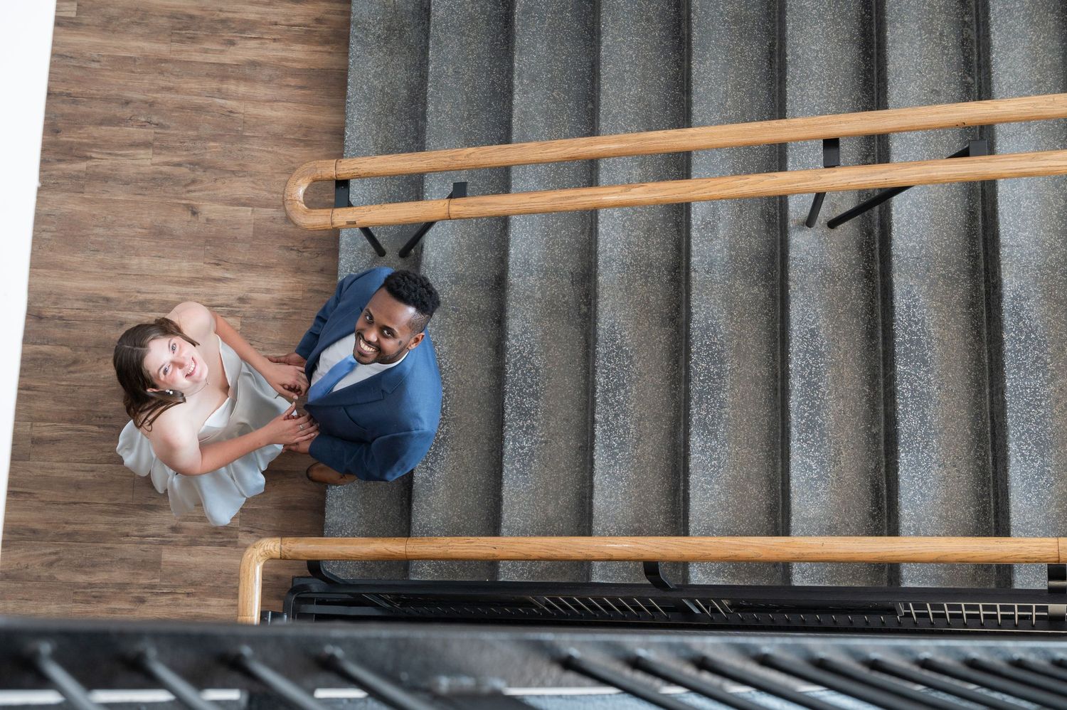 Aerial view of a man and woman smiling up at camera while walking up concrete stairs with wooden railings.