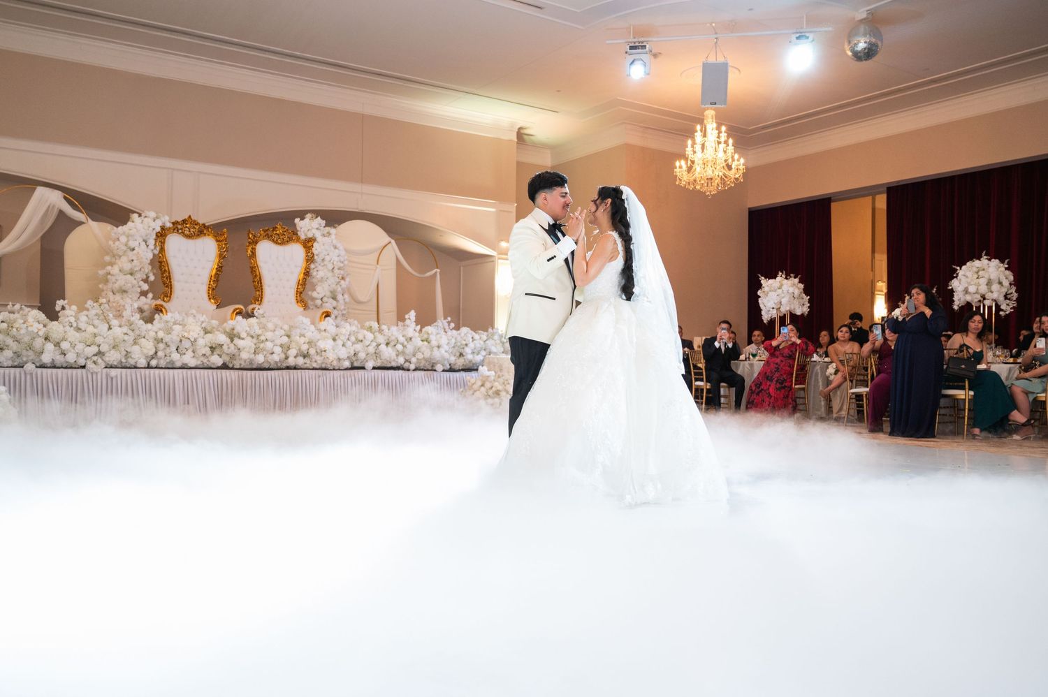 Bride and groom share first dance on white dance floor surrounded by elegant floral decor and wedding guests.