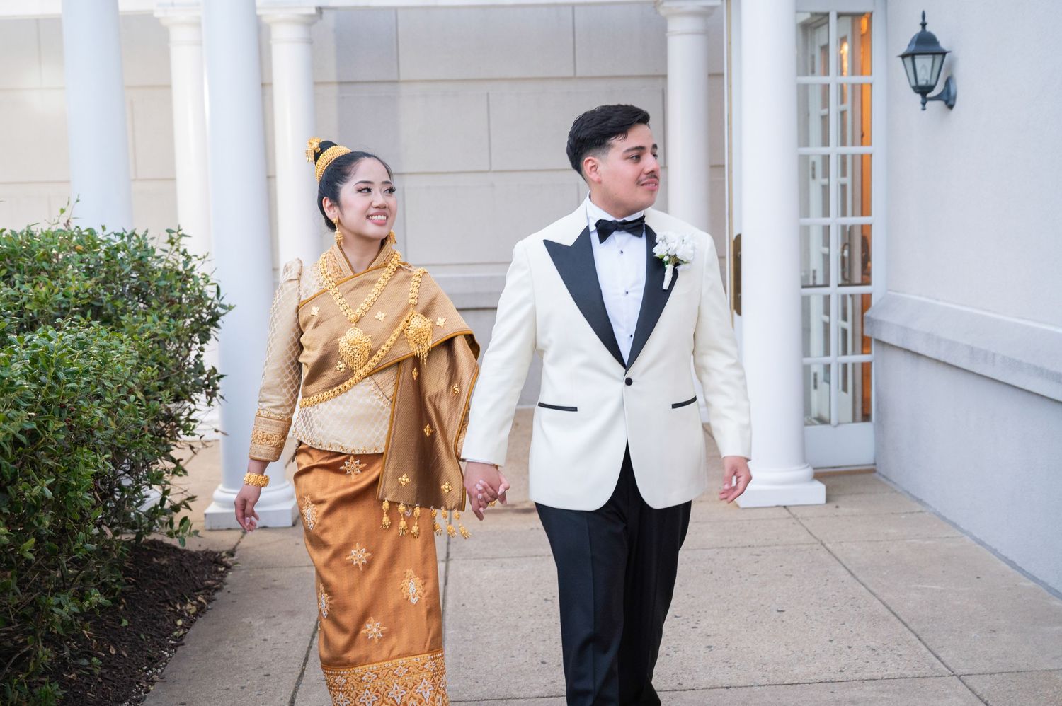 Couple in traditional Lao wedding attire and white tuxedo walking hand in hand outside elegant venue.