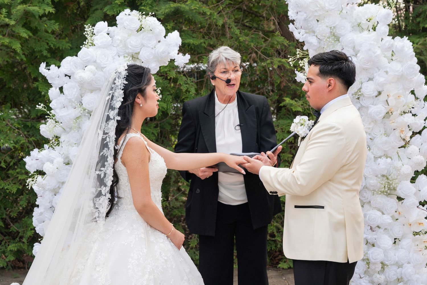 Bride and groom exchange vows at floral white arch with officiant during outdoor wedding ceremony.