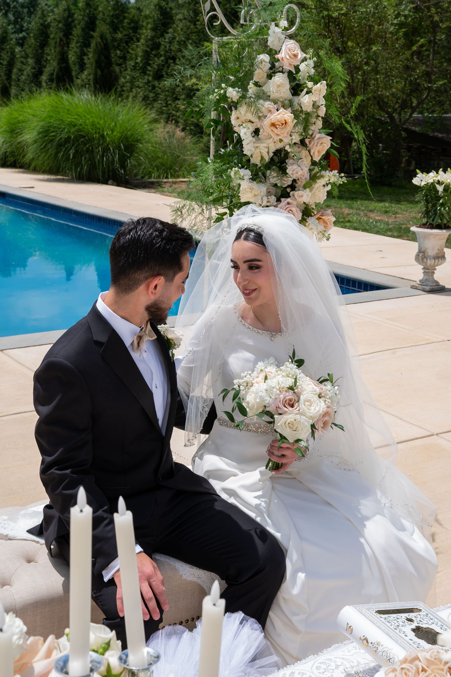 Bride and groom sitting together outdoors near a pool, with white candles and floral arrangements at their wedding ceremony.