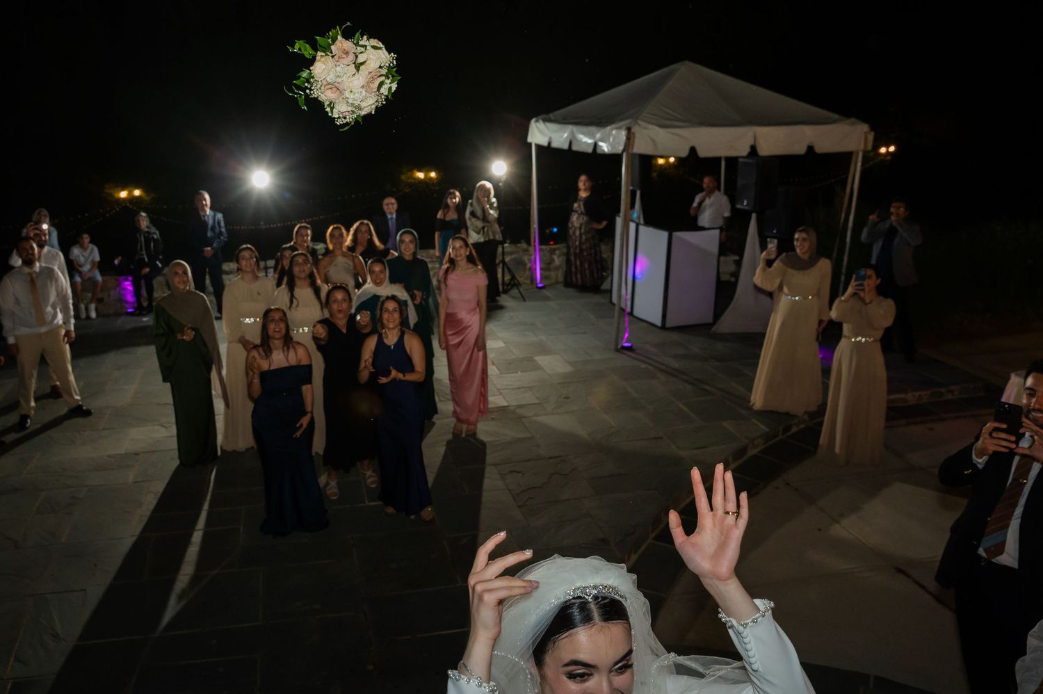Bride tossing bouquet to excited wedding guests at an outdoor nighttime reception celebration.