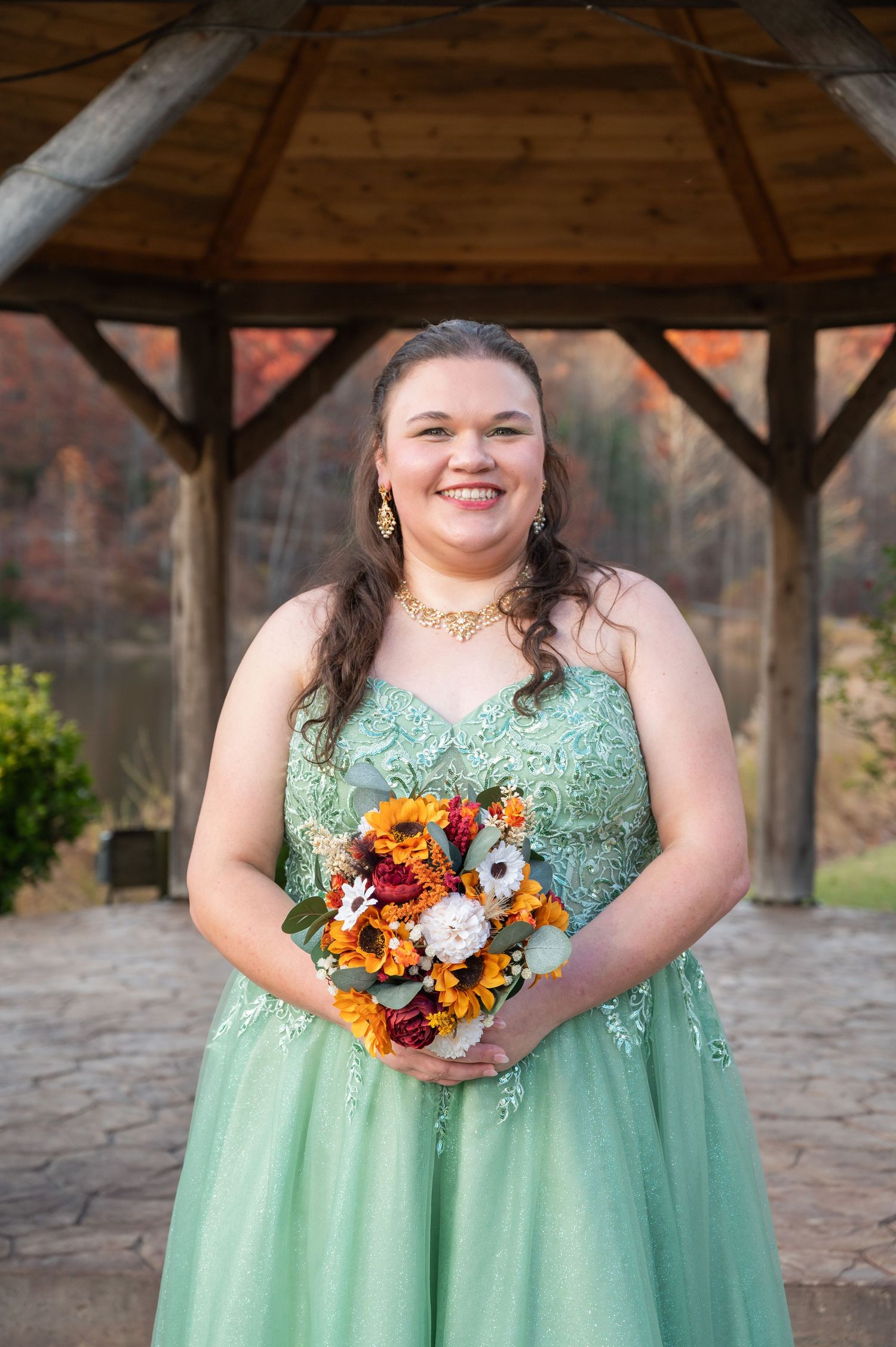Smiling woman in sage green lace gown holding autumn bouquet with sunflowers stands before wooden gazebo outdoors.