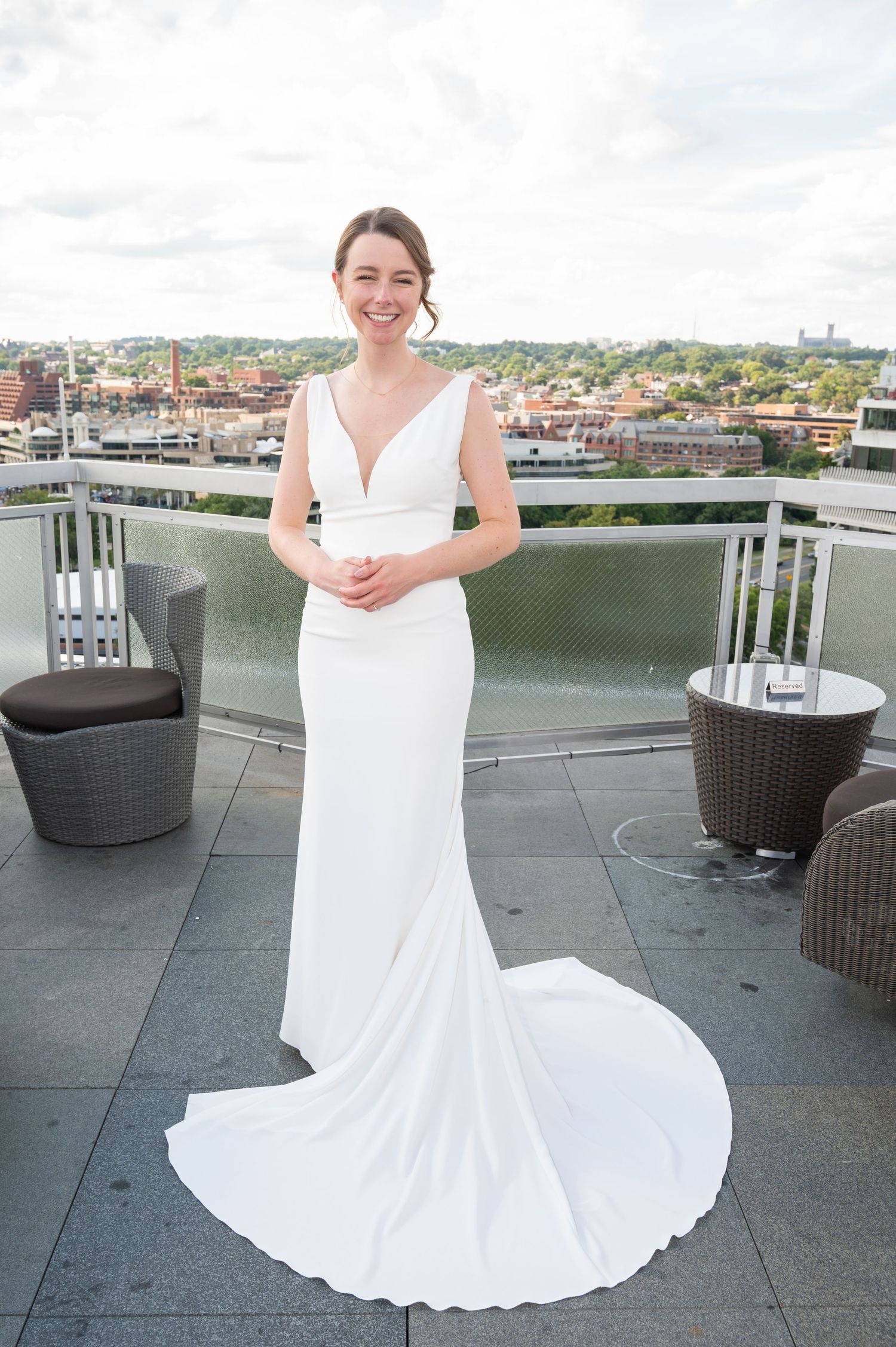 Elegant bride in sleek white V-neck gown with train posing on rooftop terrace with city views.