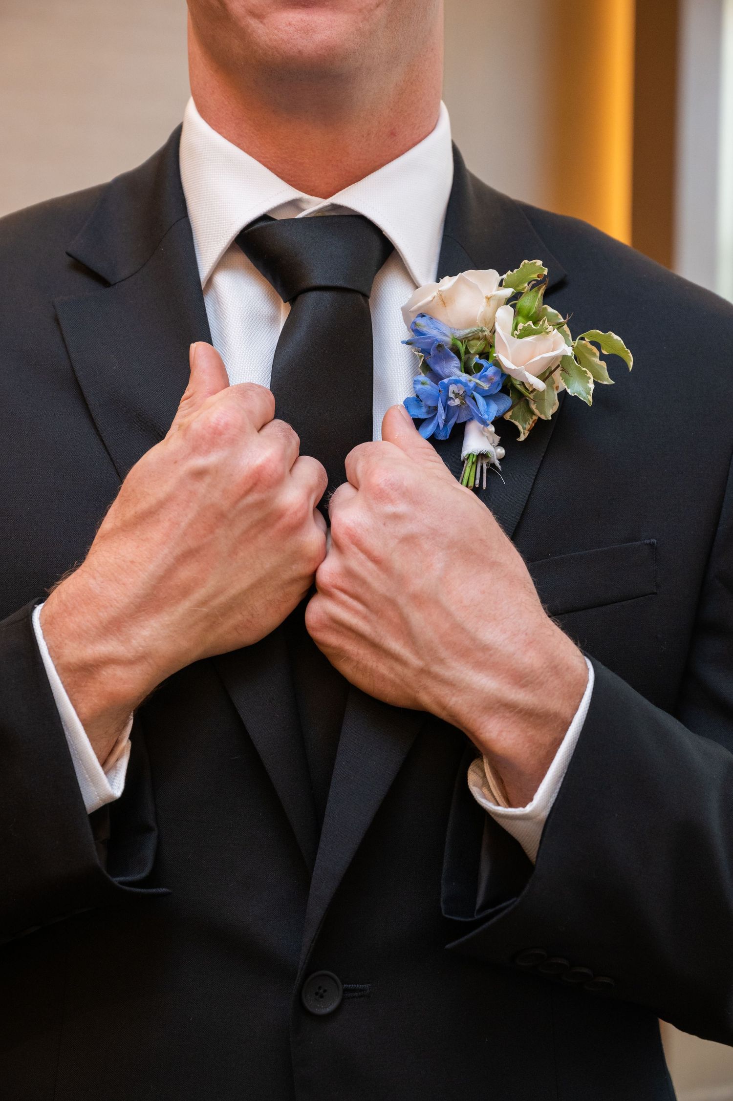 Groom in black suit adjusting tie with blue and white floral boutonniere on lapel.