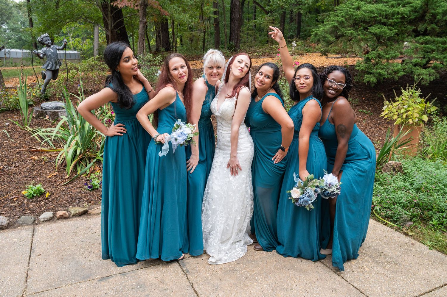 Bride posing with six bridesmaids in teal dresses in an outdoor garden wedding setting.