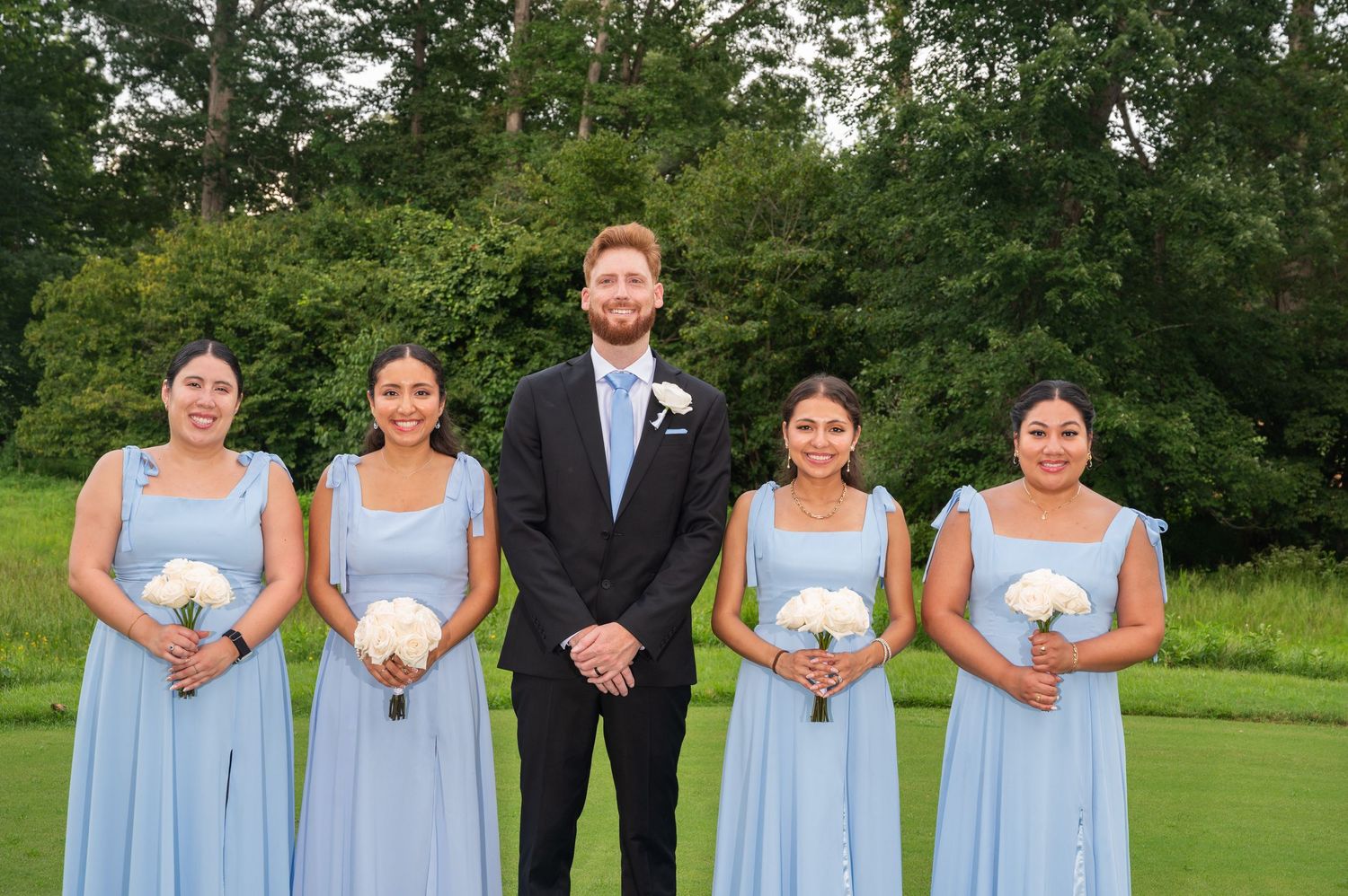 Groom in navy suit with four bridesmaids in light blue dresses holding white bouquets outdoors.
