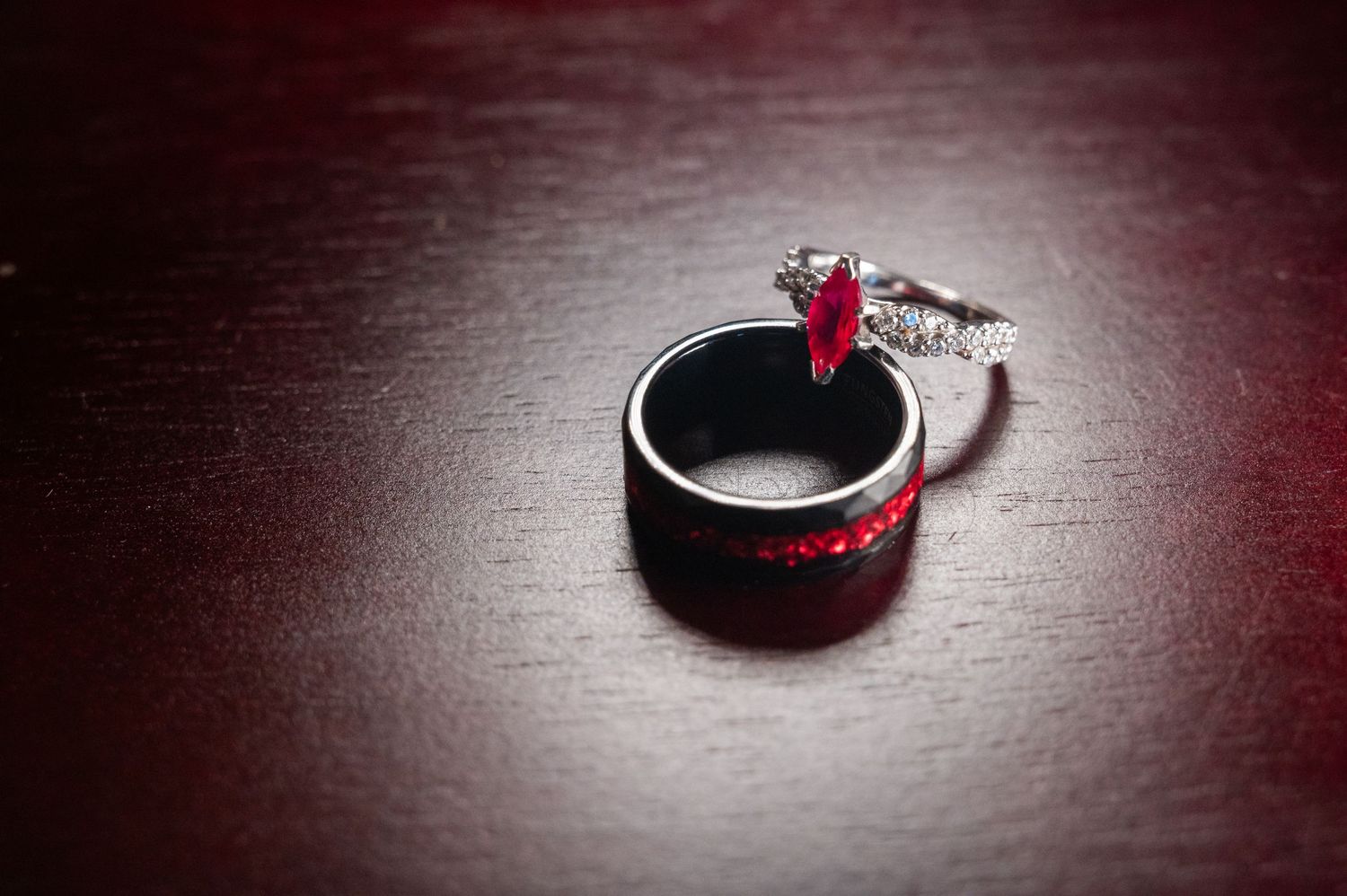 Ruby engagement ring and black wedding band resting on wooden surface.
