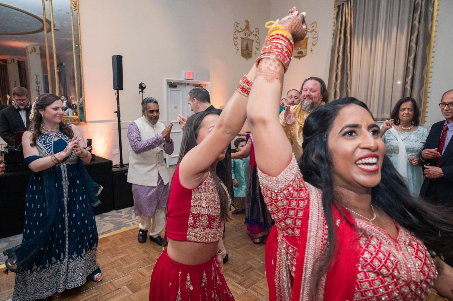 Women in red lehengas dance joyfully at a vibrant Indian wedding reception with guests celebrating in background.