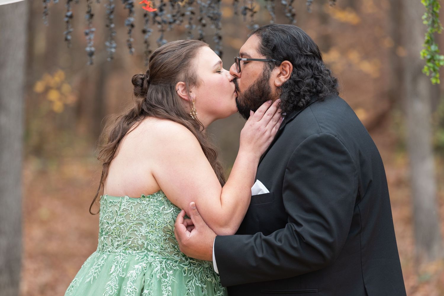 Couple sharing a romantic kiss outdoors in autumn, woman in green gown and man in black suit.