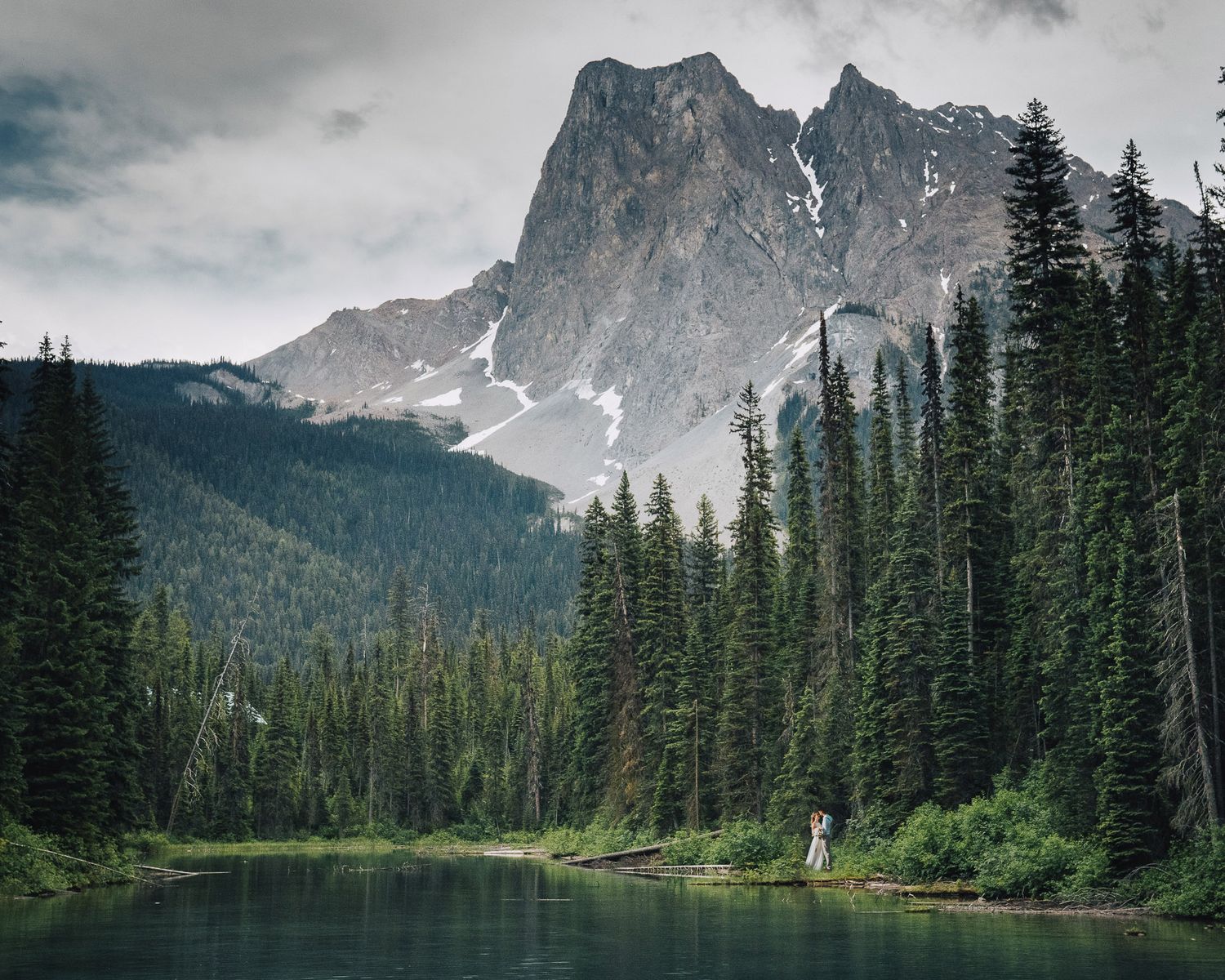 Scenic mountain lake surrounded by evergreen forest and rugged peaks under cloudy skies in wilderness landscape.