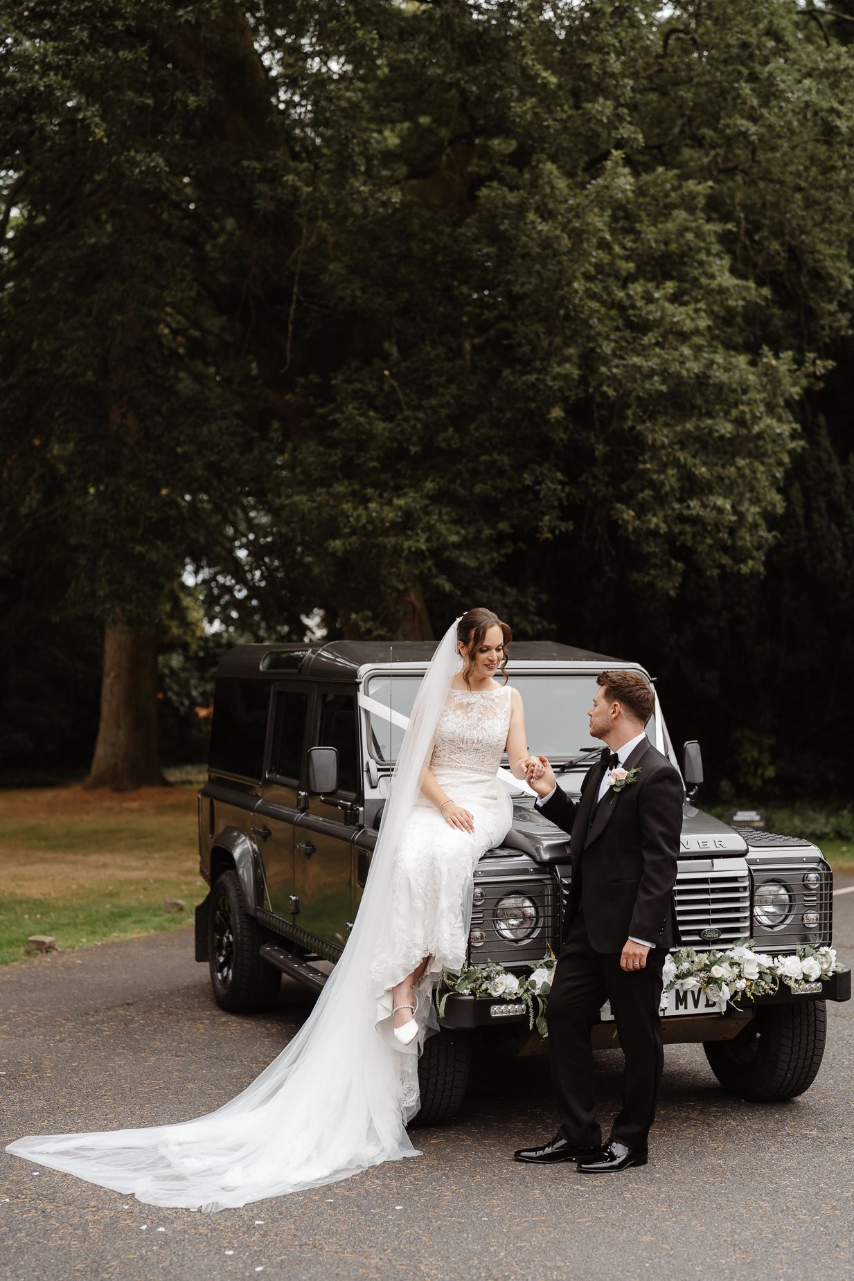 A romantic wedding scene unfolds in front of a vintage black car as a couple shares a tender moment in a wooded setting.