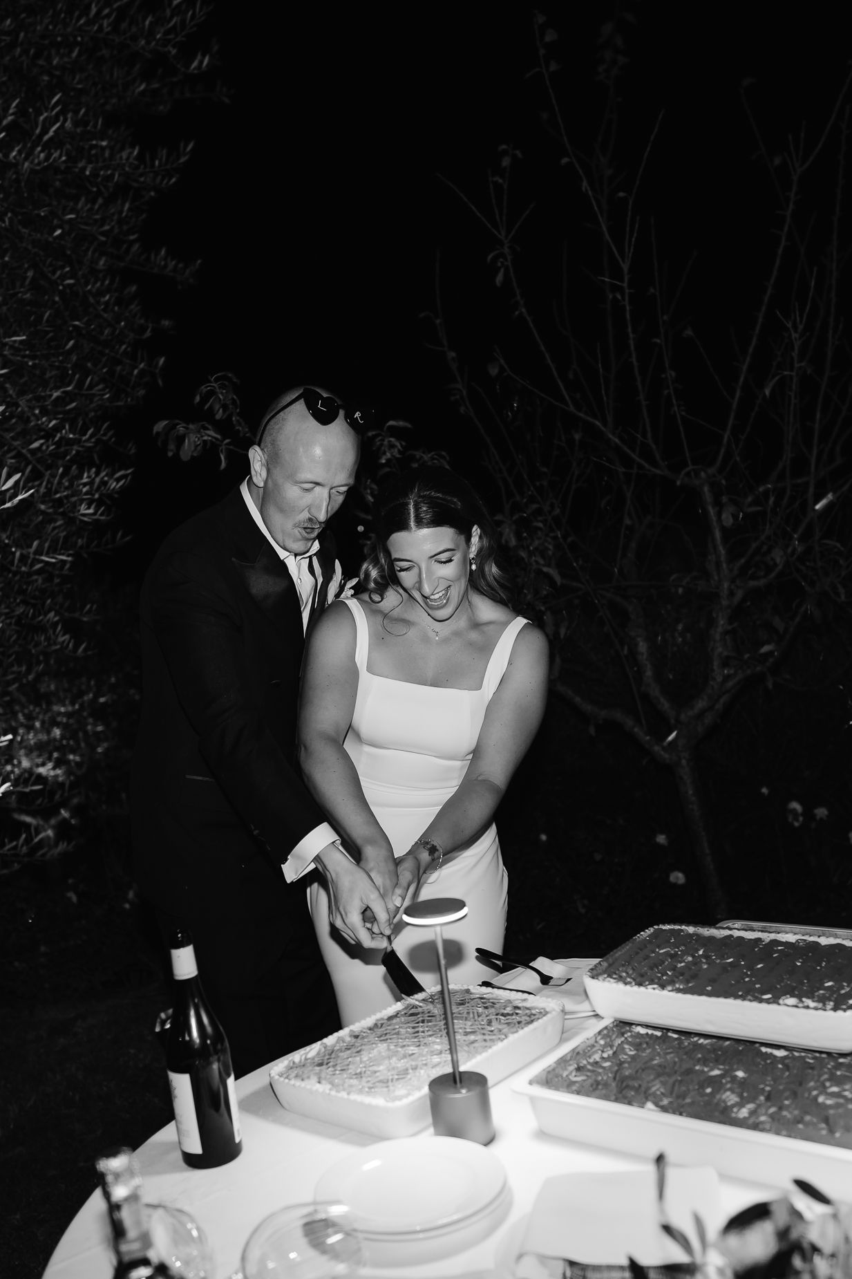 A black and white photo of a couple cutting their wedding cake at night.