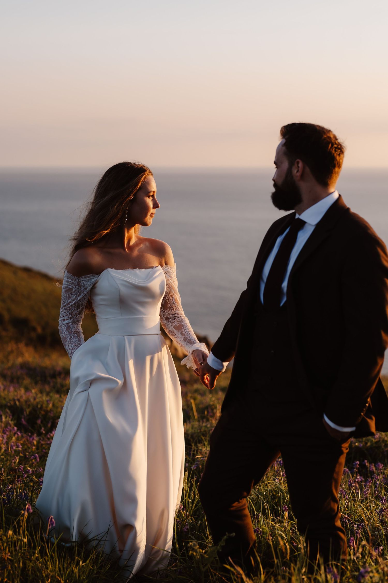 A couple in wedding attire walks through a scenic coastal meadow at sunset, creating romantic silhouettes against the ocean.
