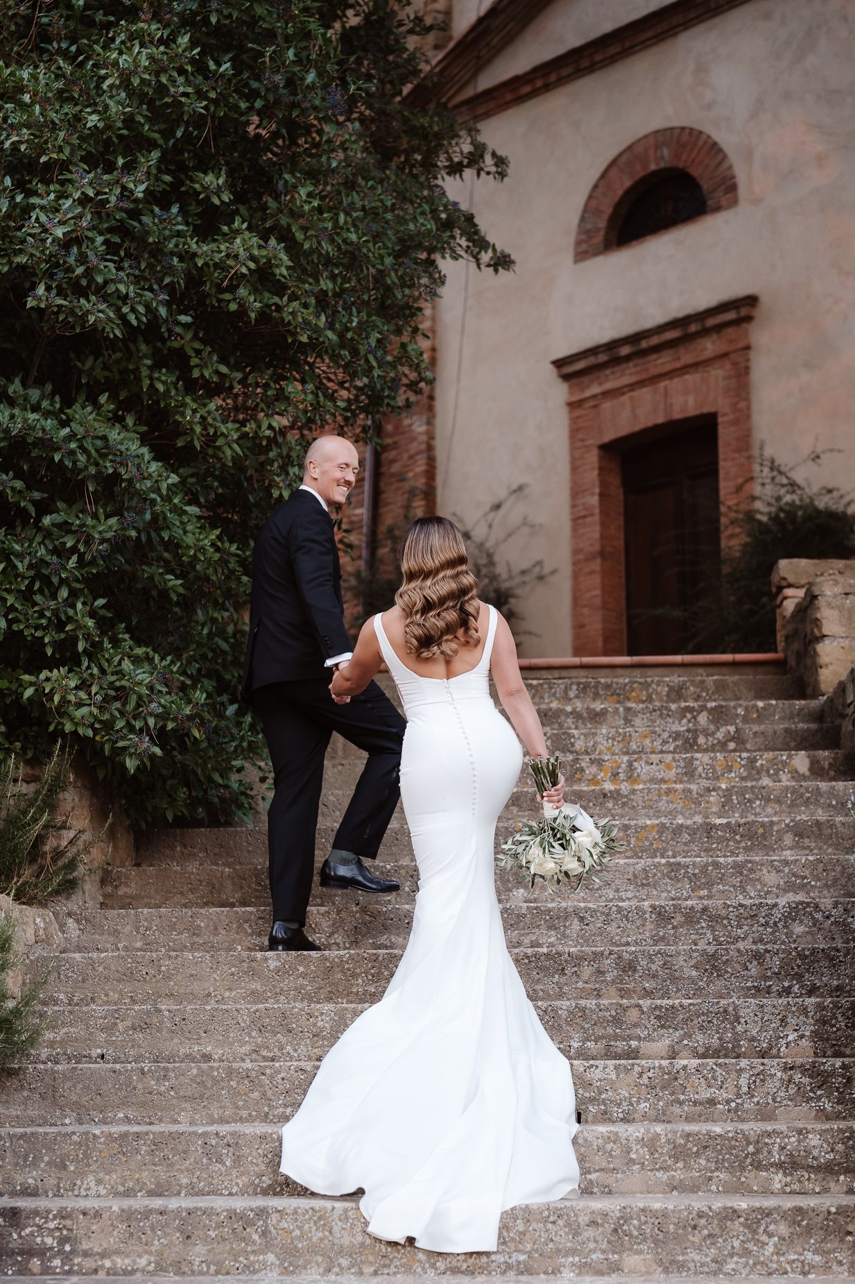 A couple in wedding attire climbing stone steps at a historic church entrance with red brick archways.