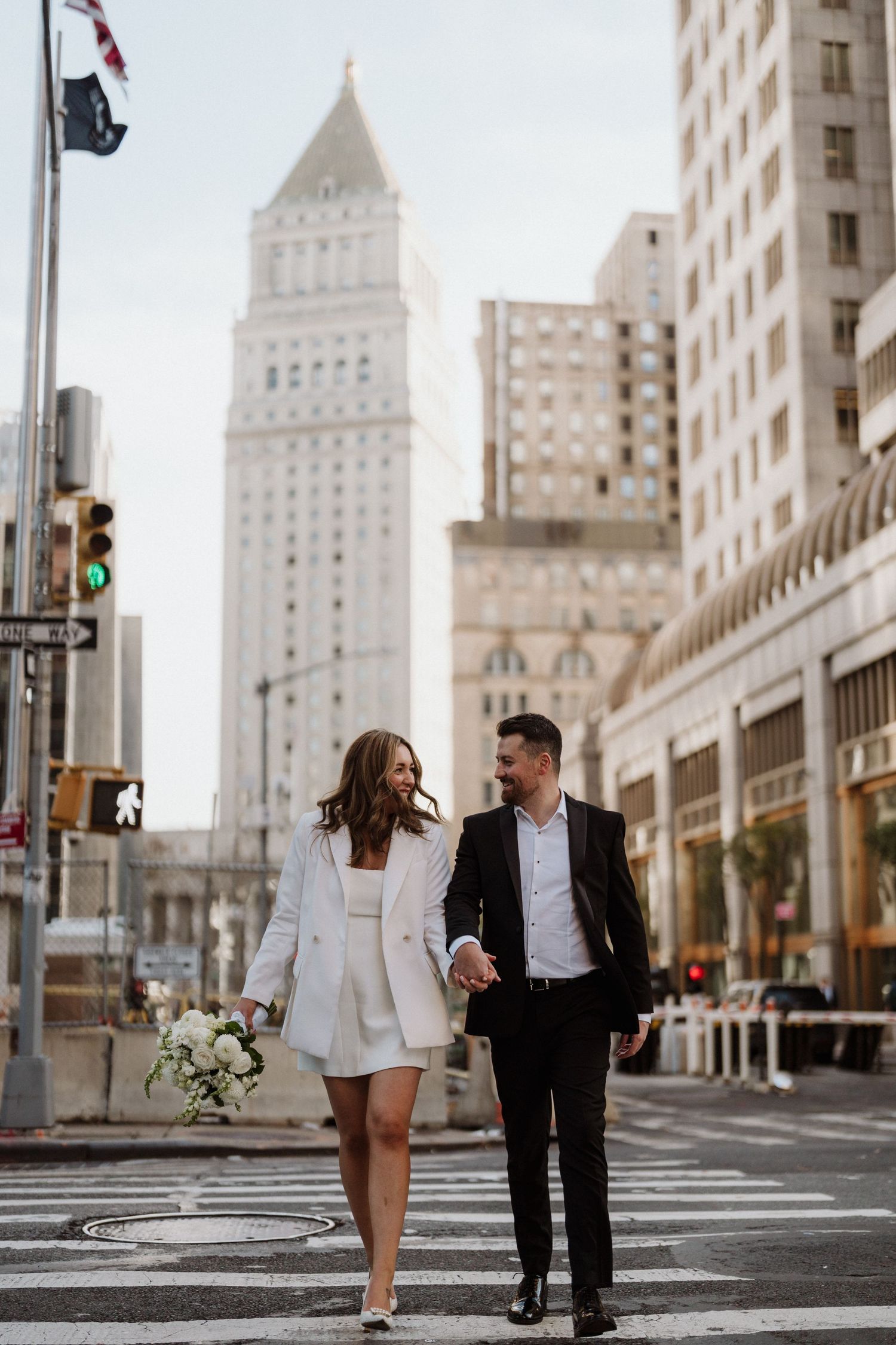 A couple walks hand in hand across a city street with tall art deco buildings in the background.