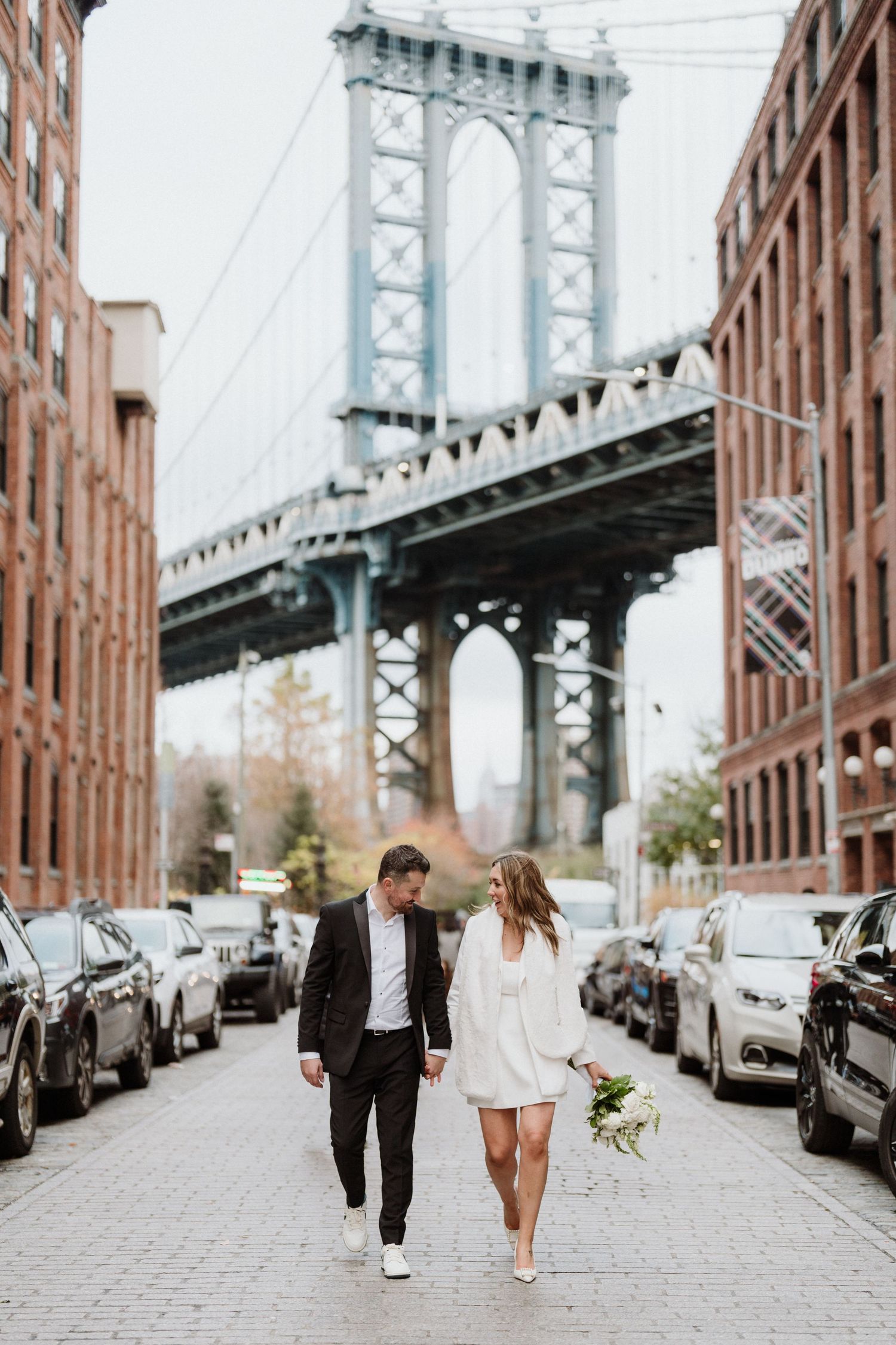 A couple walks along a cobblestone street with the Manhattan Bridge in the background in DUMBO, Brooklyn.