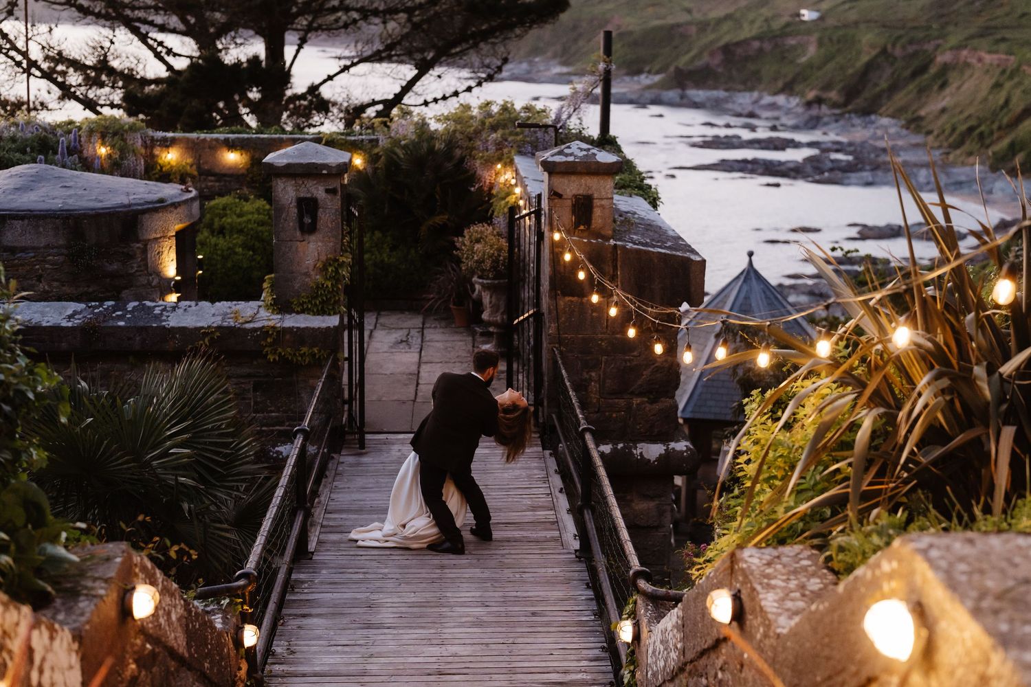 String lights illuminate a cozy wooden deck overlooking the ocean at sunset during an intimate outdoor gathering.