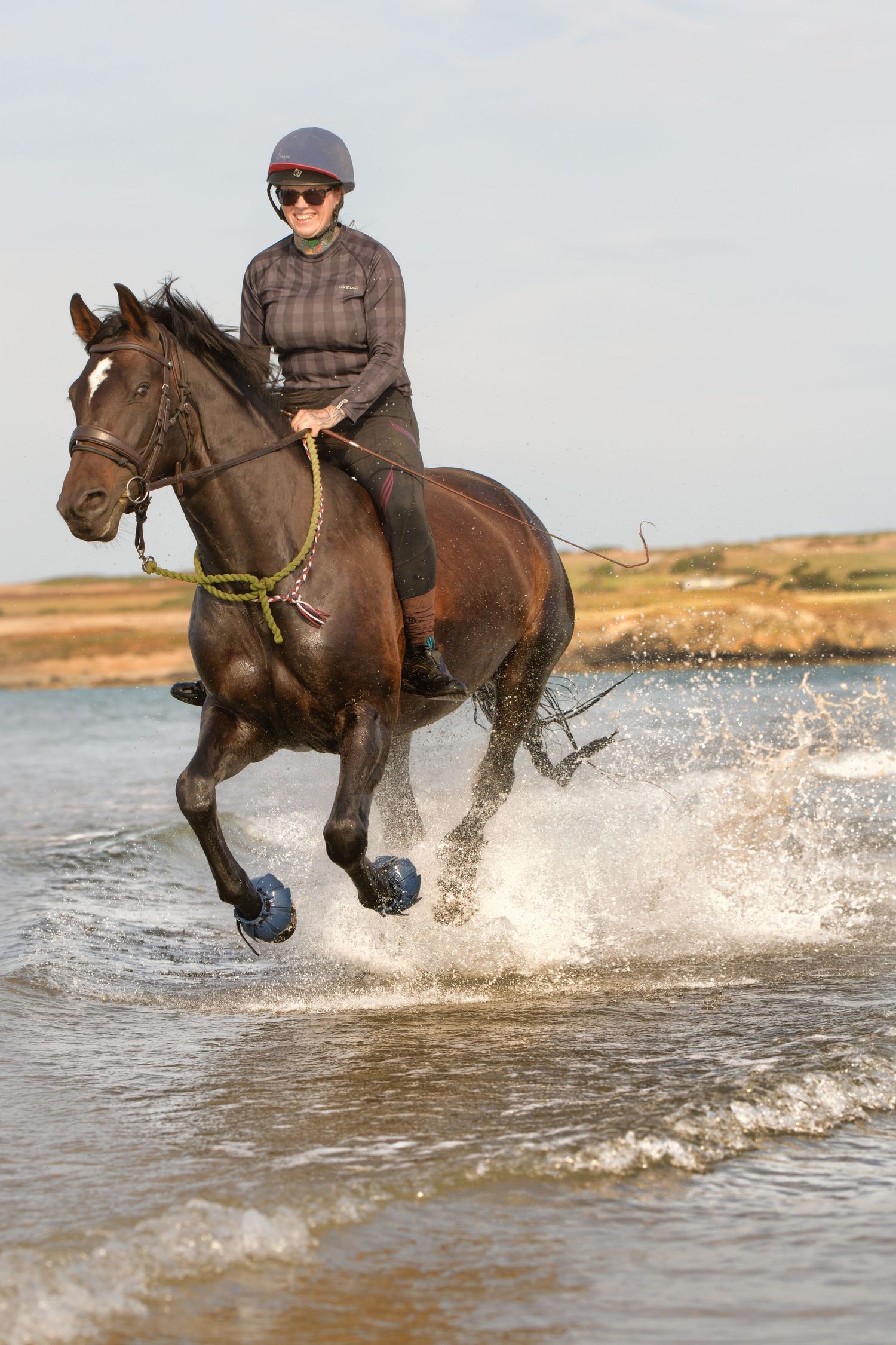 Galloping horse splashing through waves on the beach under a bright blue sky, rider smiling with joy during an equine photography shoot