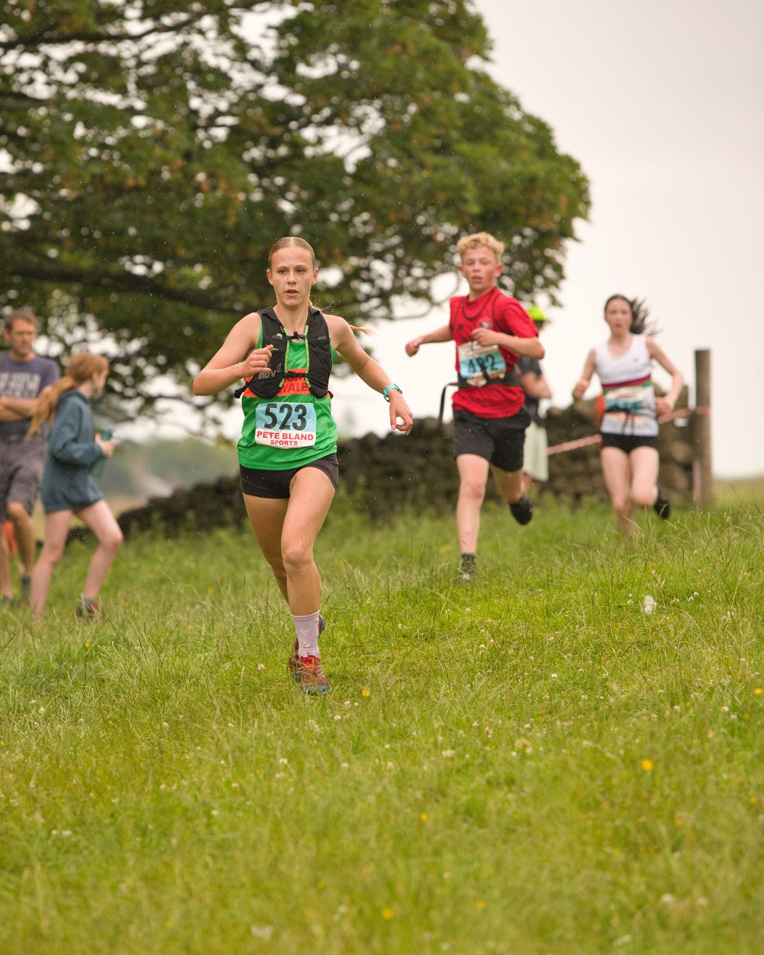 Junior fell runners descending a steep grassy hillside during a race, focused and moving fast with mountains in the background.
