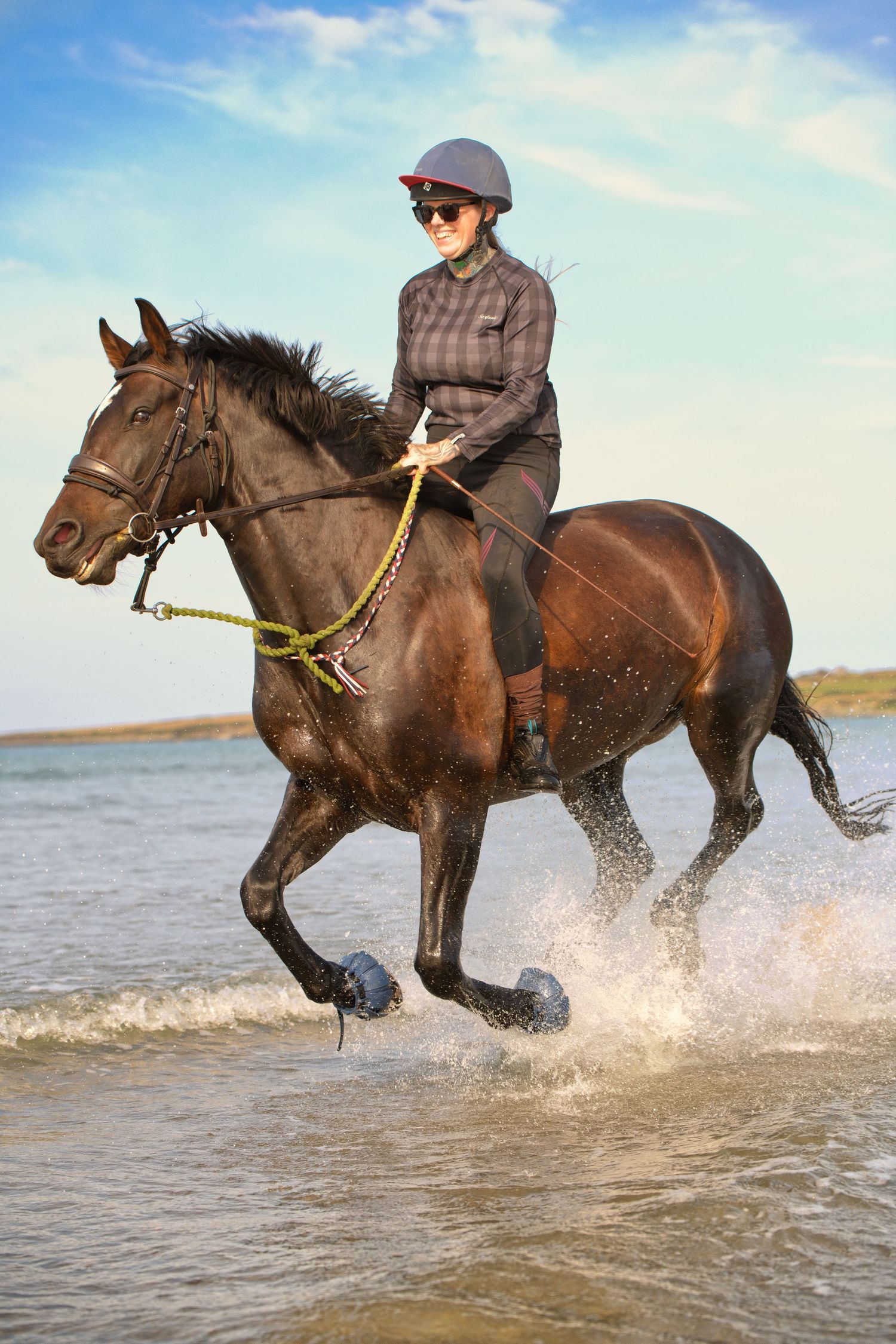 Galloping horse splashing through waves on the beach under a bright blue sky, rider smiling with joy during an equine photography shoot