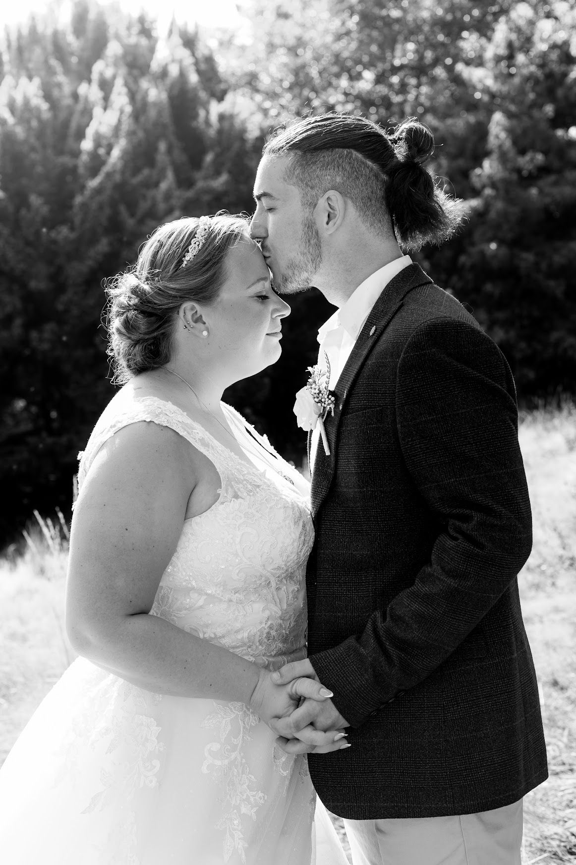 Black and white photo of groom with man bun kissing brides forehead, holding hands outdoors.