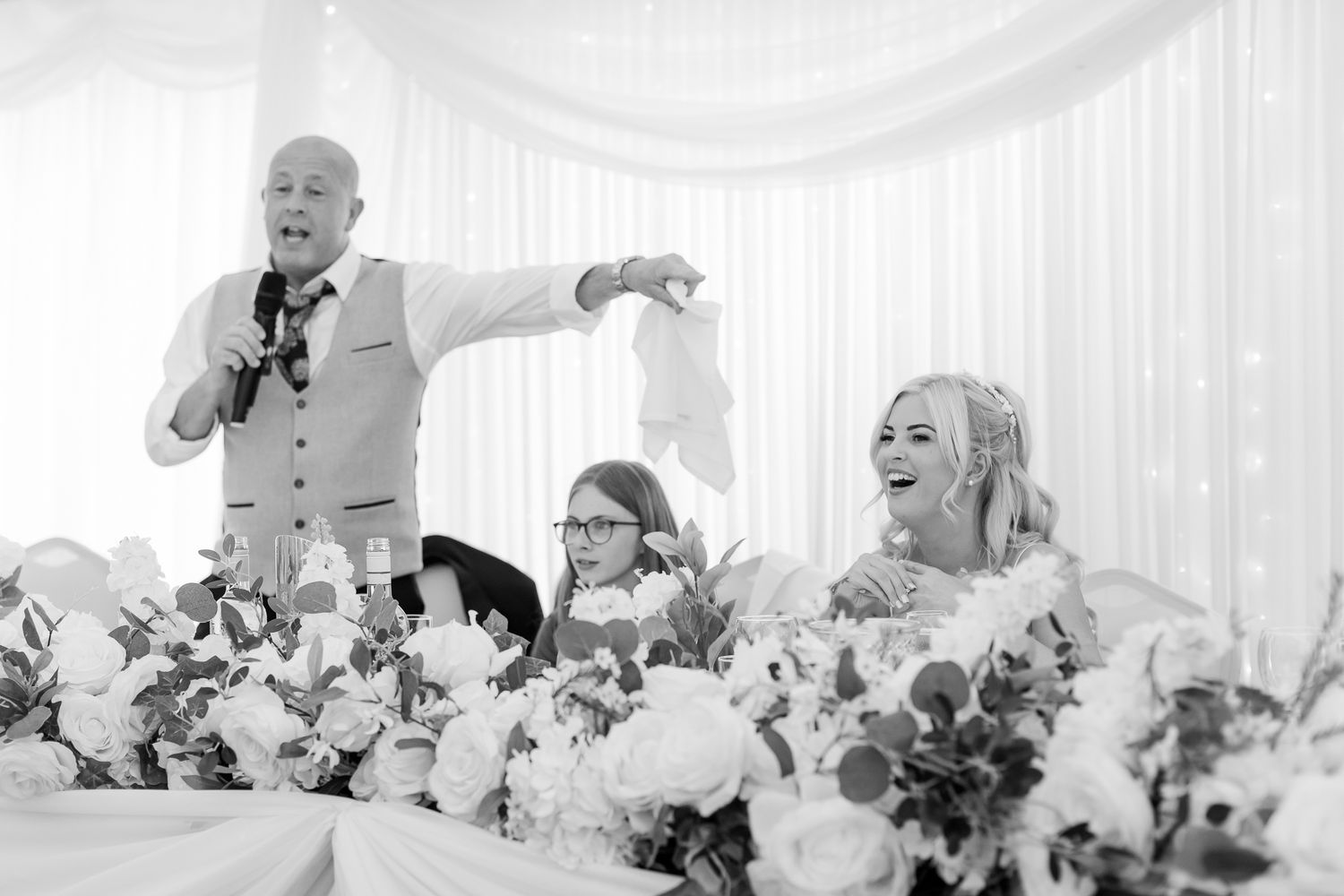 Father of the bride delivers a humorous speech as the bride laughs at the wedding reception.
