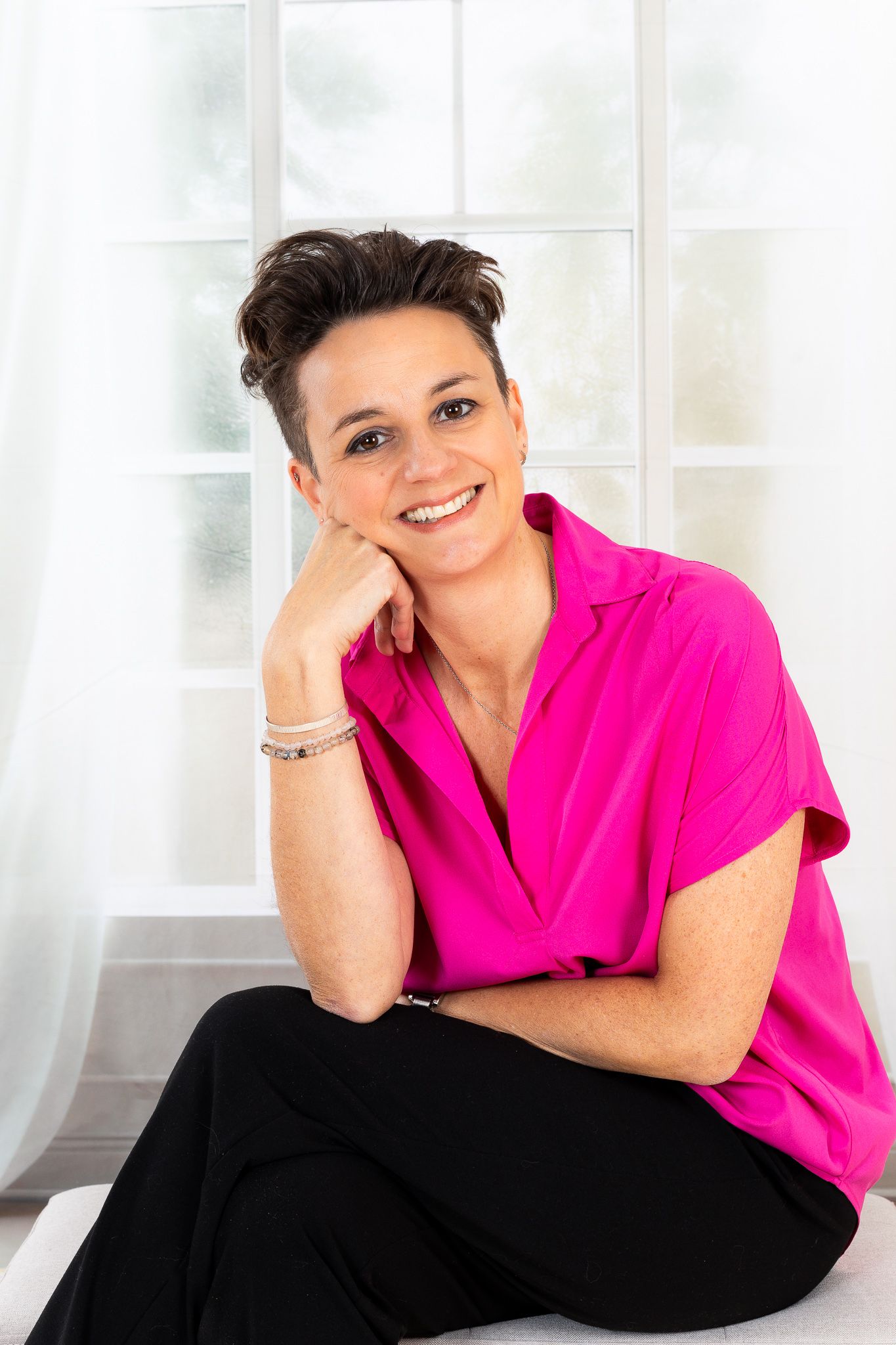 Cheerful short-haired woman in a pink blouse sitting and smiling near a bright window.