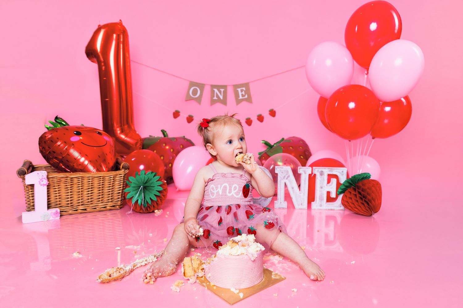 Baby girl celebrates first birthday smash cake with strawberry theme, pink balloons, and ONE decorations on pink background.