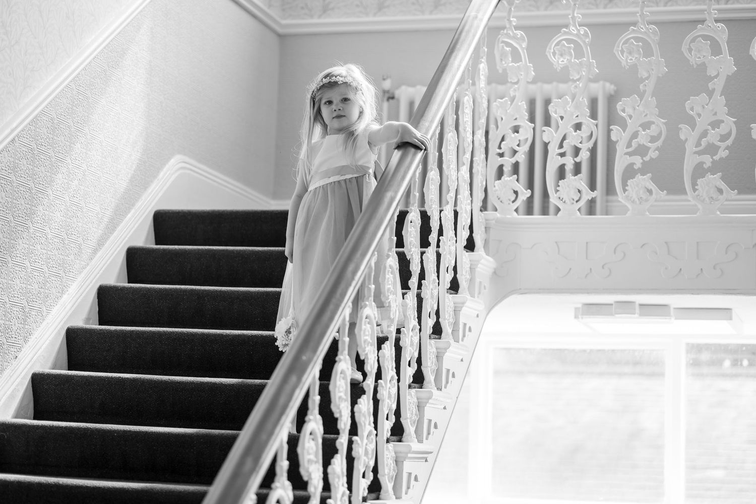 Young girl in white dress standing on elegant staircase with ornate white railings, black and white photography.