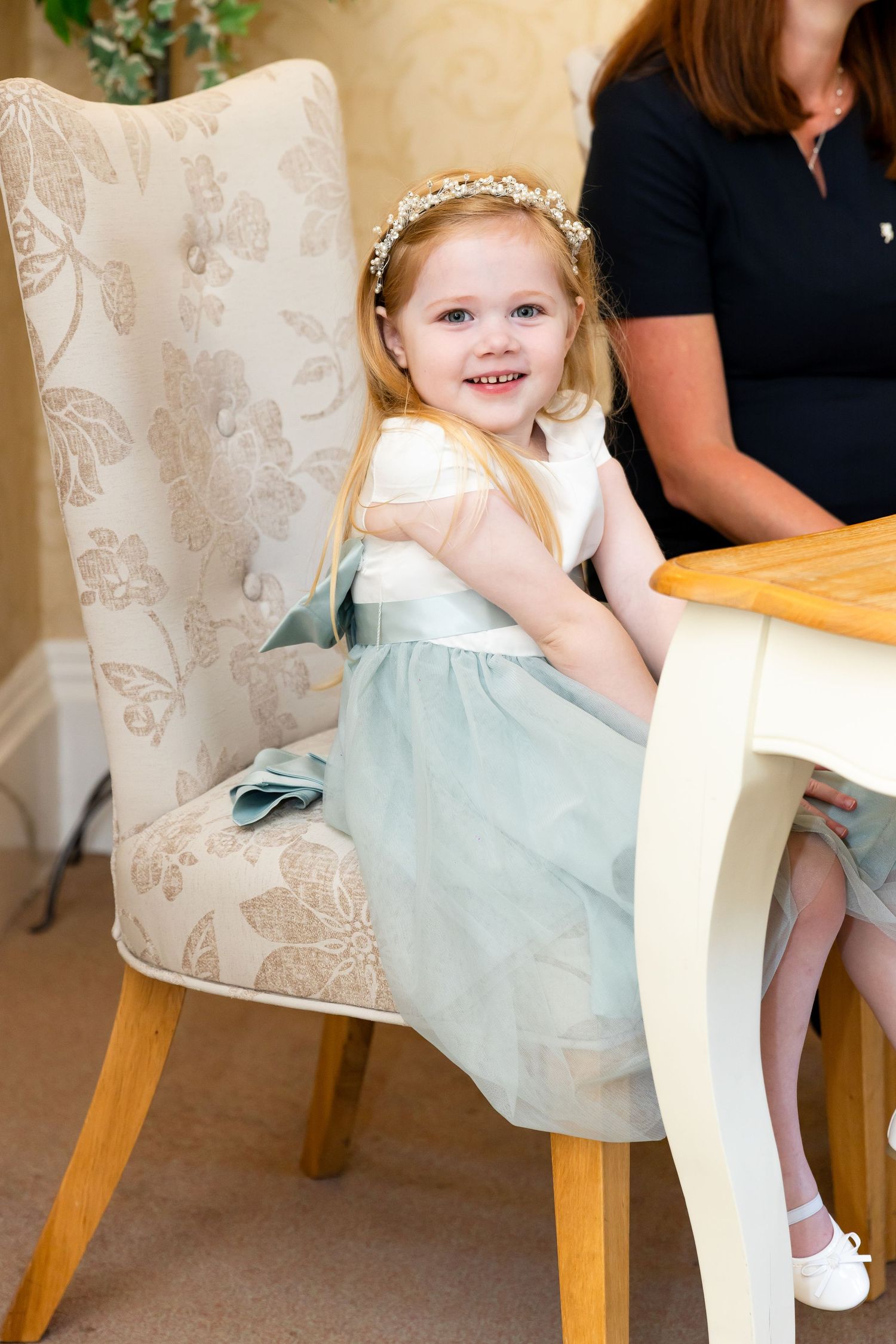 Smiling little flower girl in mint tulle dress and floral crown sitting at a table during a wedding celebration.