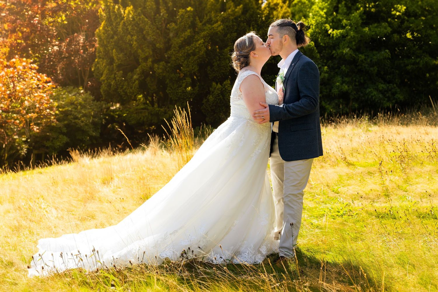 Bride and groom kissing in golden sunlight in a meadow, bride wearing a long white gown.